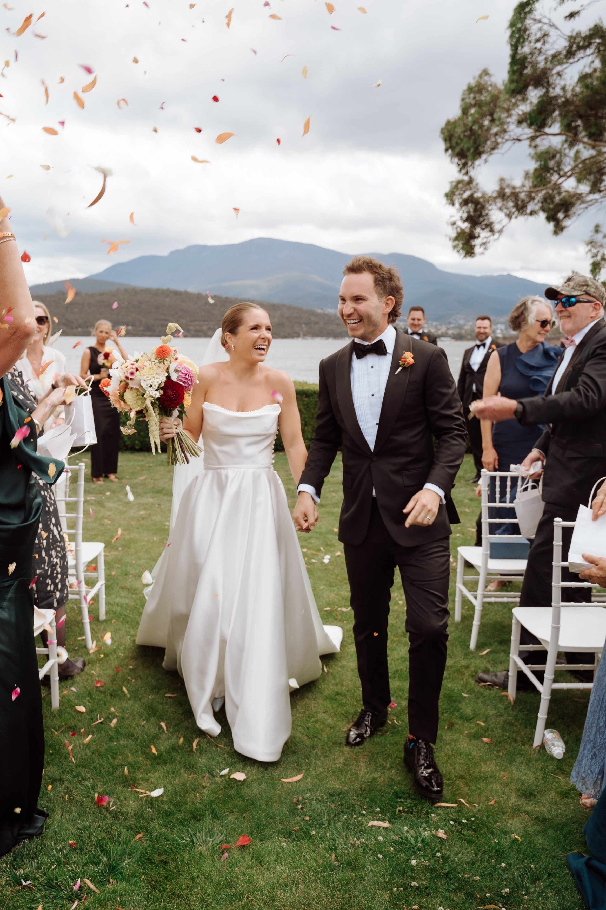 A newly married couple walking hand-in-hand outdoors during their wedding, surrounded by guests throwing flower petals, with a lake, mountains, and cloudy sky in the background.