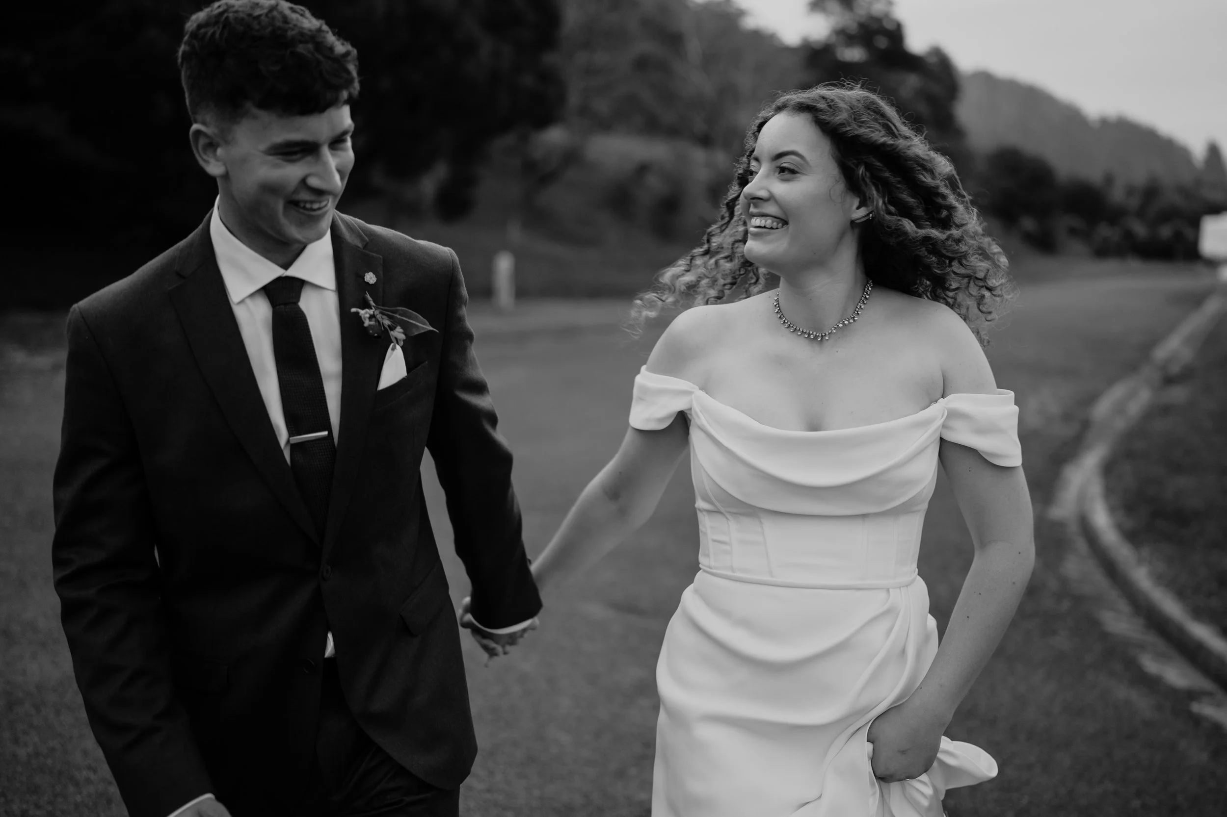 A black and white photo of a smiling bride and groom holding hands, walking outdoors in a scenic area.