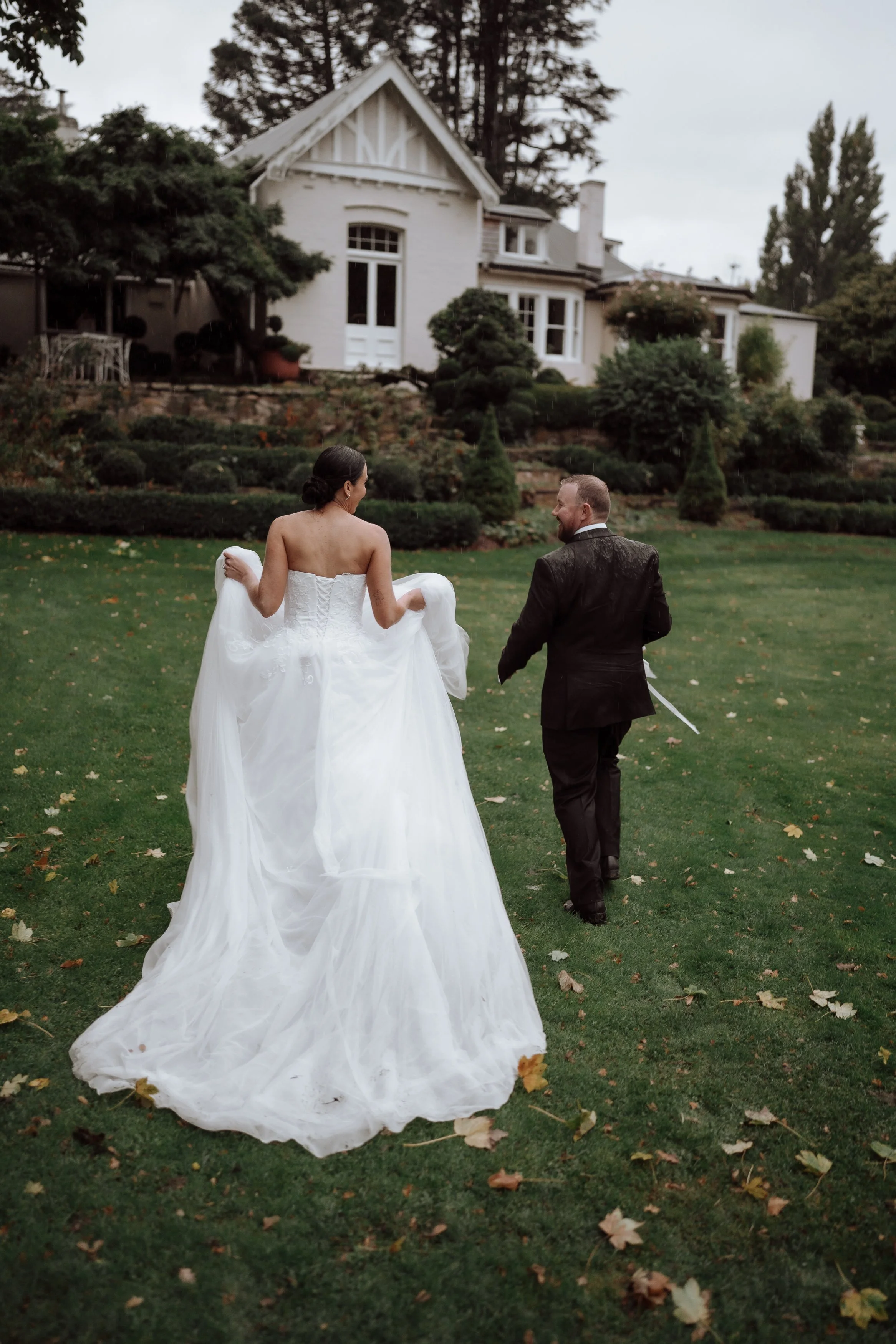 A bride and groom walking on a grassy backyard lawn near a large white house with trees and bushes.