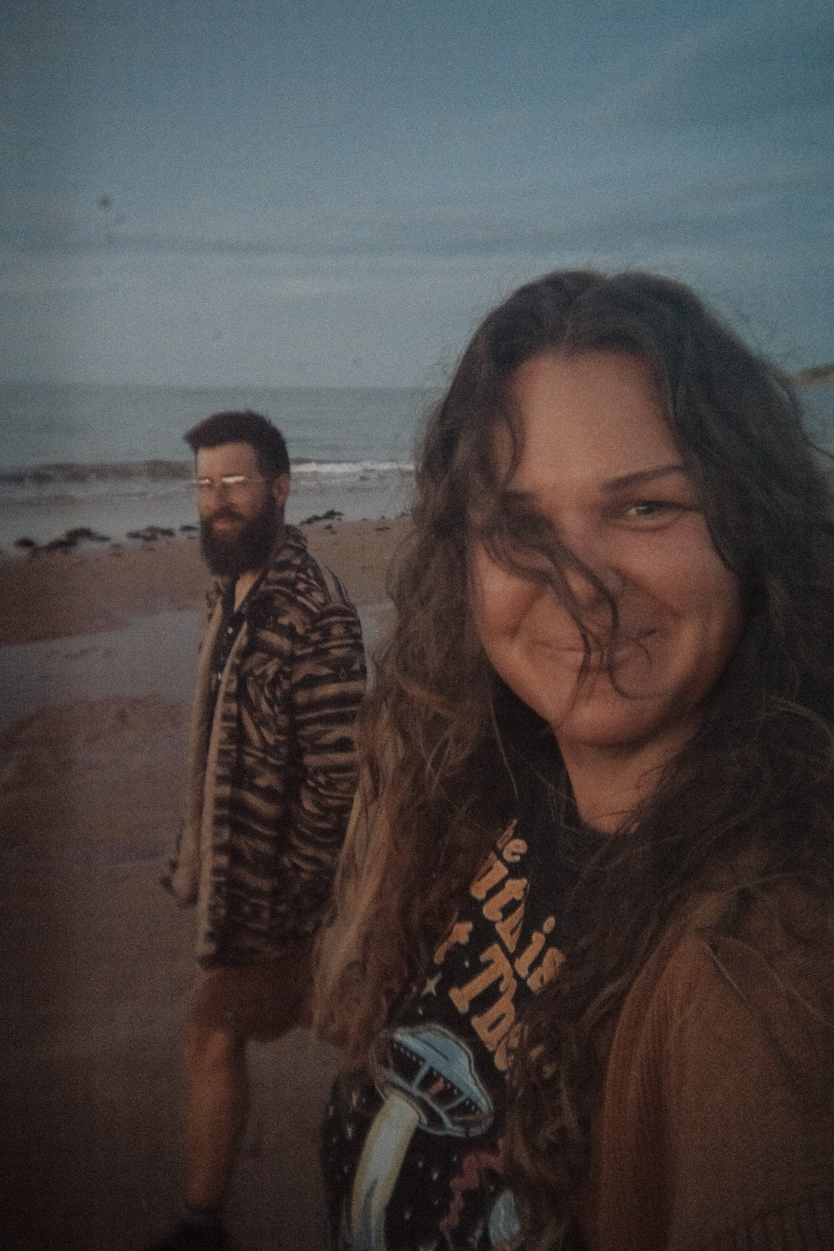 A woman smiling in the foreground, and a man with a beard and glasses in the background, standing on a beach at dusk or dawn with the ocean and cloudy sky behind them.