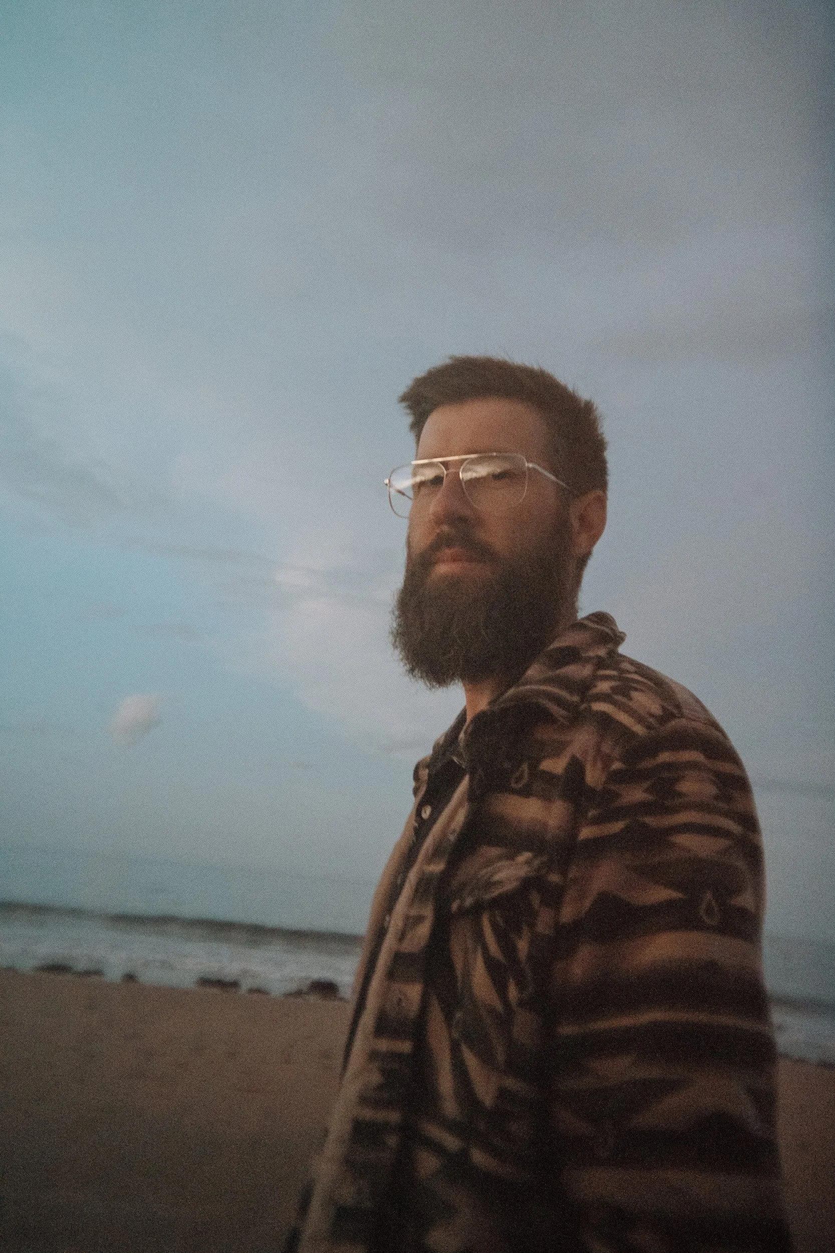 A man with glasses, a full beard, and a patterned shirt looks over his shoulder on a beach during dusk. He's thinking about photographing luxury wedding in Tasmania