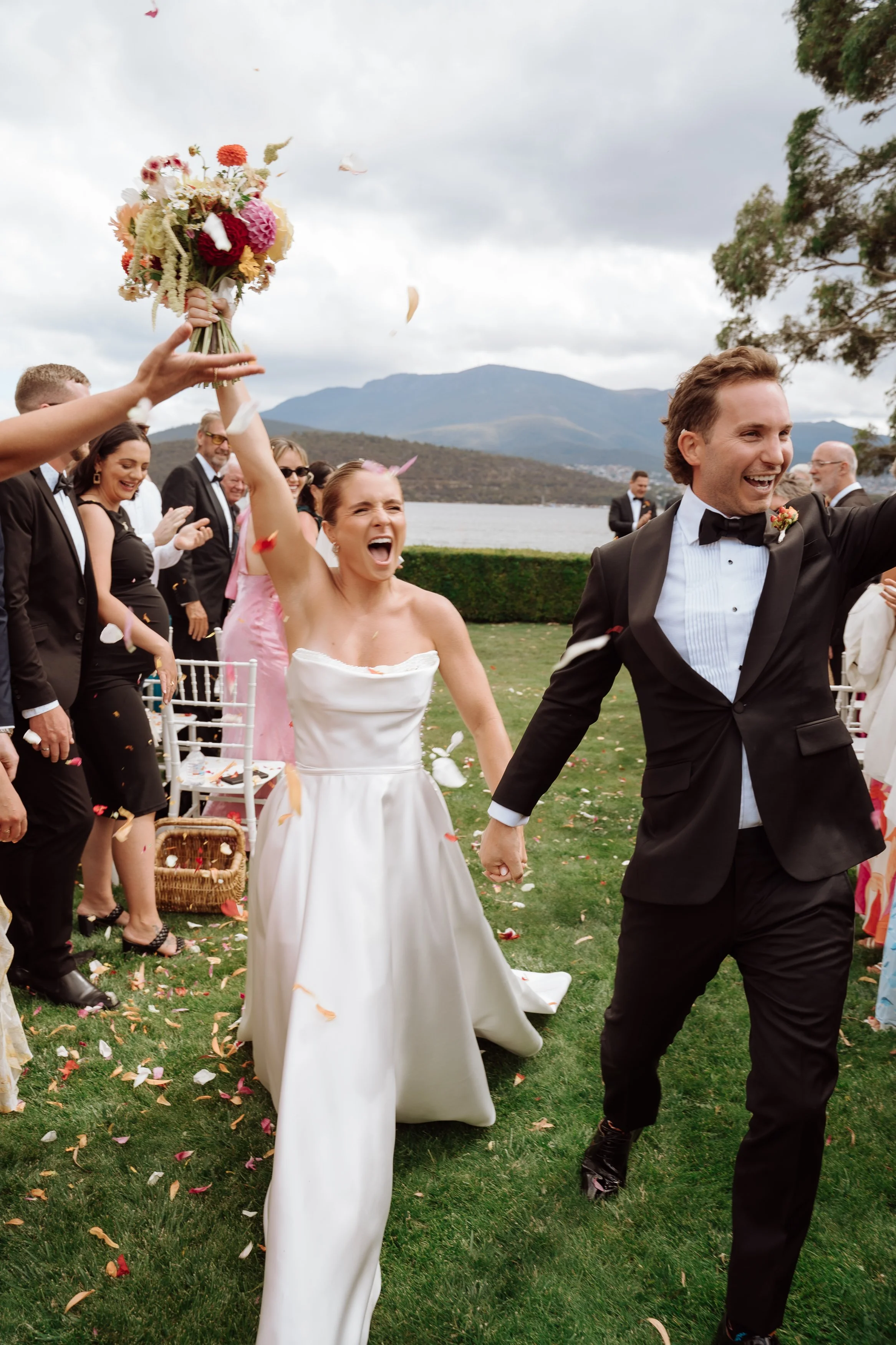 Bridal couple celebrating outdoors at a wedding ceremony. The bride is holding a bouquet and cheering with her mouth open, while the groom is happy and smiling. Guests are clapping and throwing confetti on a grassy area near a lake with mountains in the background.