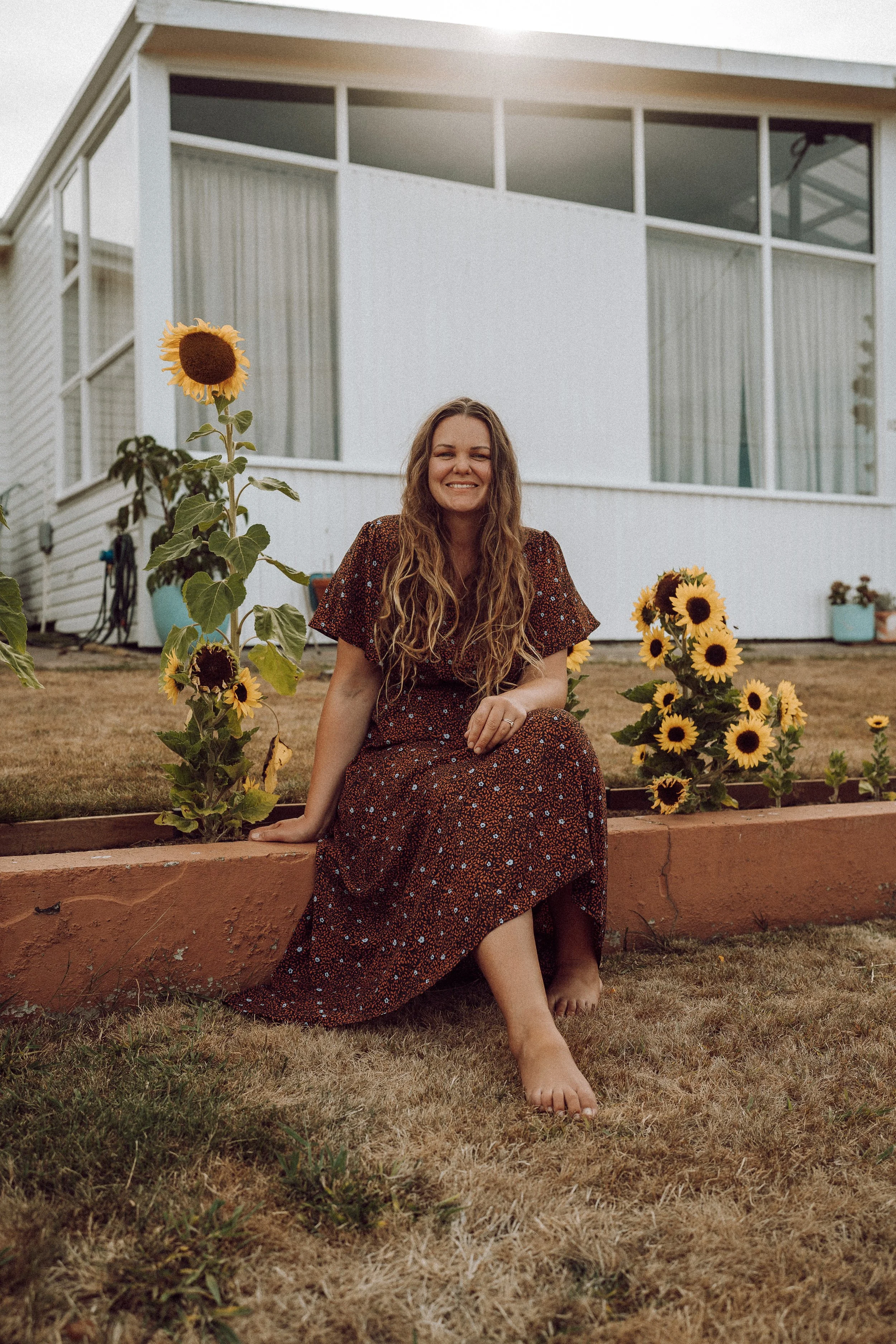 A woman in a floral dress sitting on a brick garden border, smiling at the camera, surrounded by sunflowers, with a white house in the background. She's thinking about photographing candid weddings in Tasmania