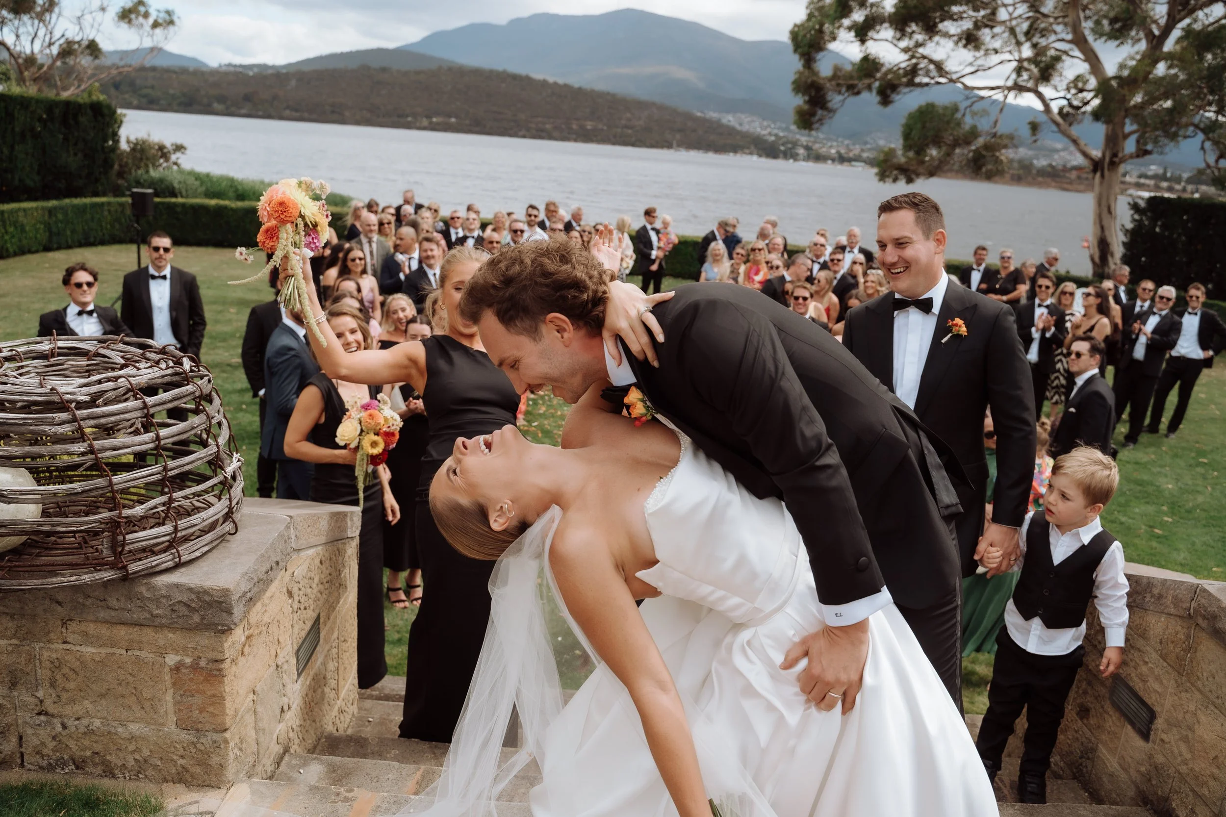 A bride and groom sharing a kiss at an outdoor wedding ceremony by a lake with mountains in the background, surrounded by wedding guests.