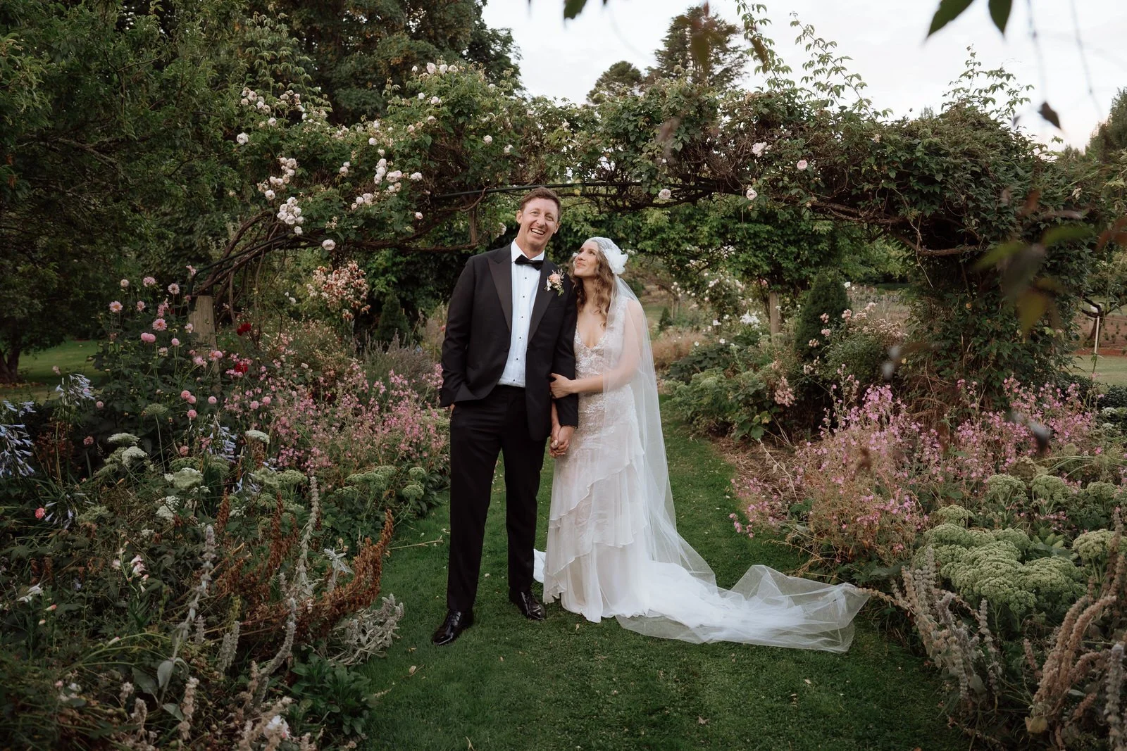 A newlywed couple standing hand-in-hand in a lush garden, smiling and gazing at each other under a flowering arch, surrounded by various colorful flowers and greenery.