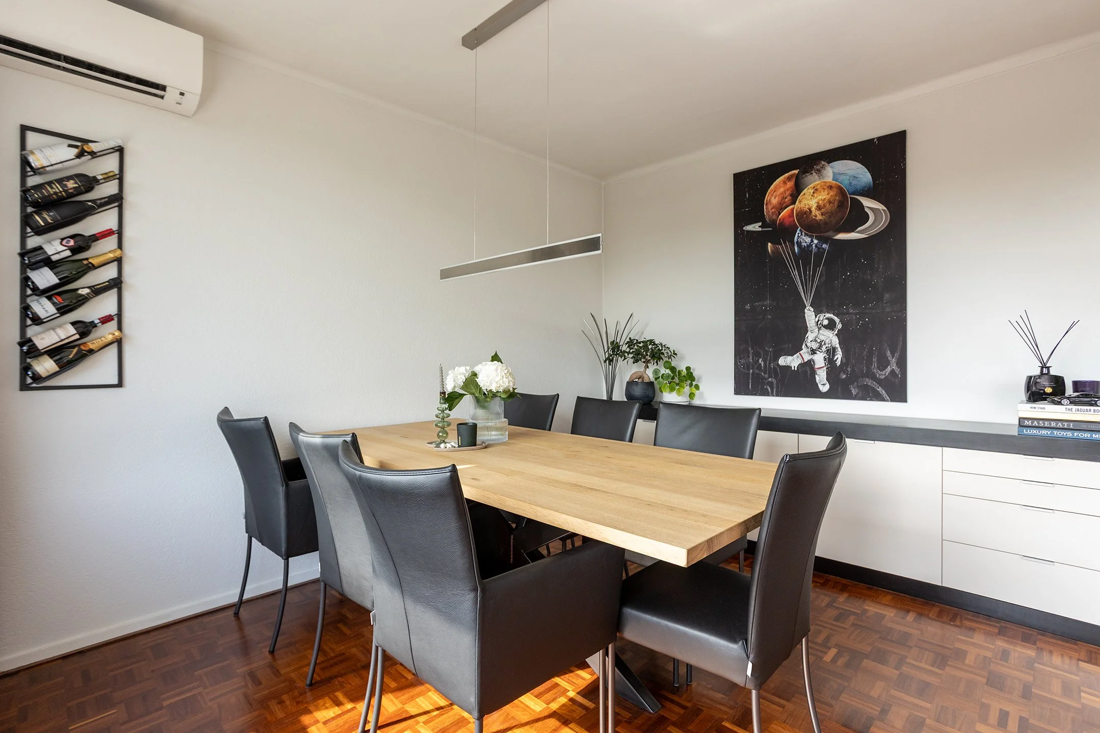 Modern diningkamer met houten tafel, zes zwart leren stoelen, planten, bloemen, muurdecoratie met planeten, en een whiteboard met boeken op de rand.