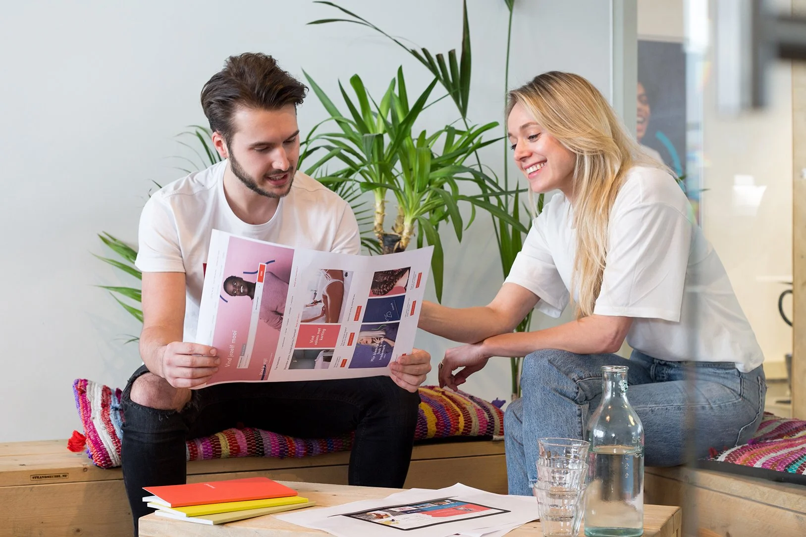 Twee jonge mensen zitten samen op een bank, kijken naar een brochure, lachen en praten. Er staat een fles water en twee glazen op de tafel.