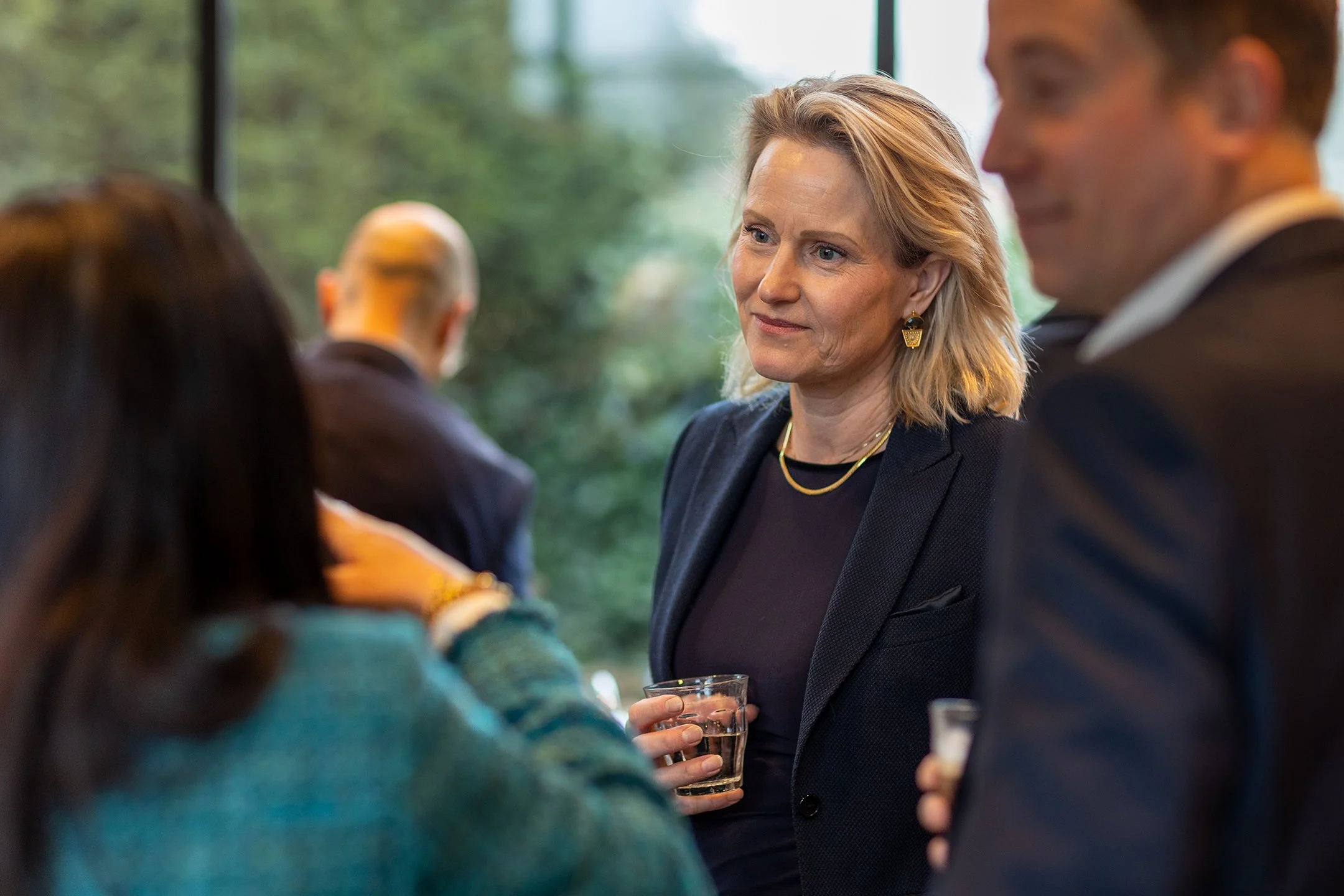 Mensen die in een groepsgesprek staan tijdens een zakelijke bijeenkomst of netwerkfeest, met een vrouw in een zwarte outfit en goudkleurige sieraden die een glas drinkt, binnen met grote ramen en groene buitenomgeving.