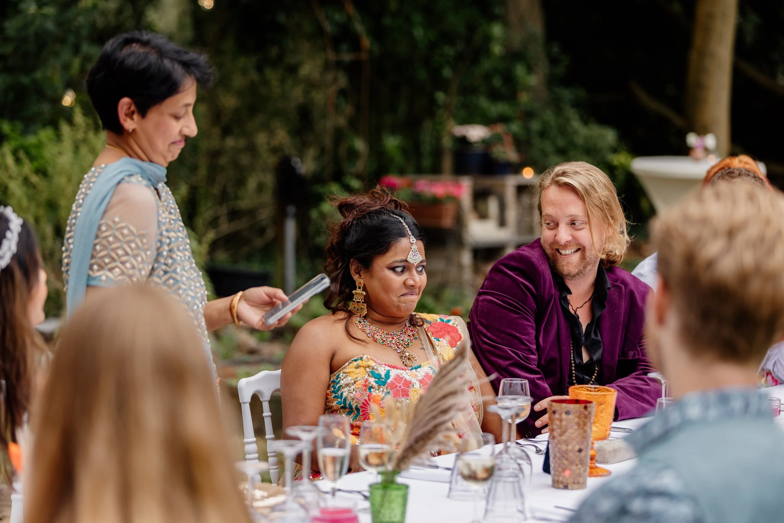 Een groep mensen zit aan een feestelijke tafel buiten, met kleurrijke decoraties en bloemen. Eén vrouw in traditionele Indiase kleding kijkt met een waarblijf, terwijl een man naast haar lacht.