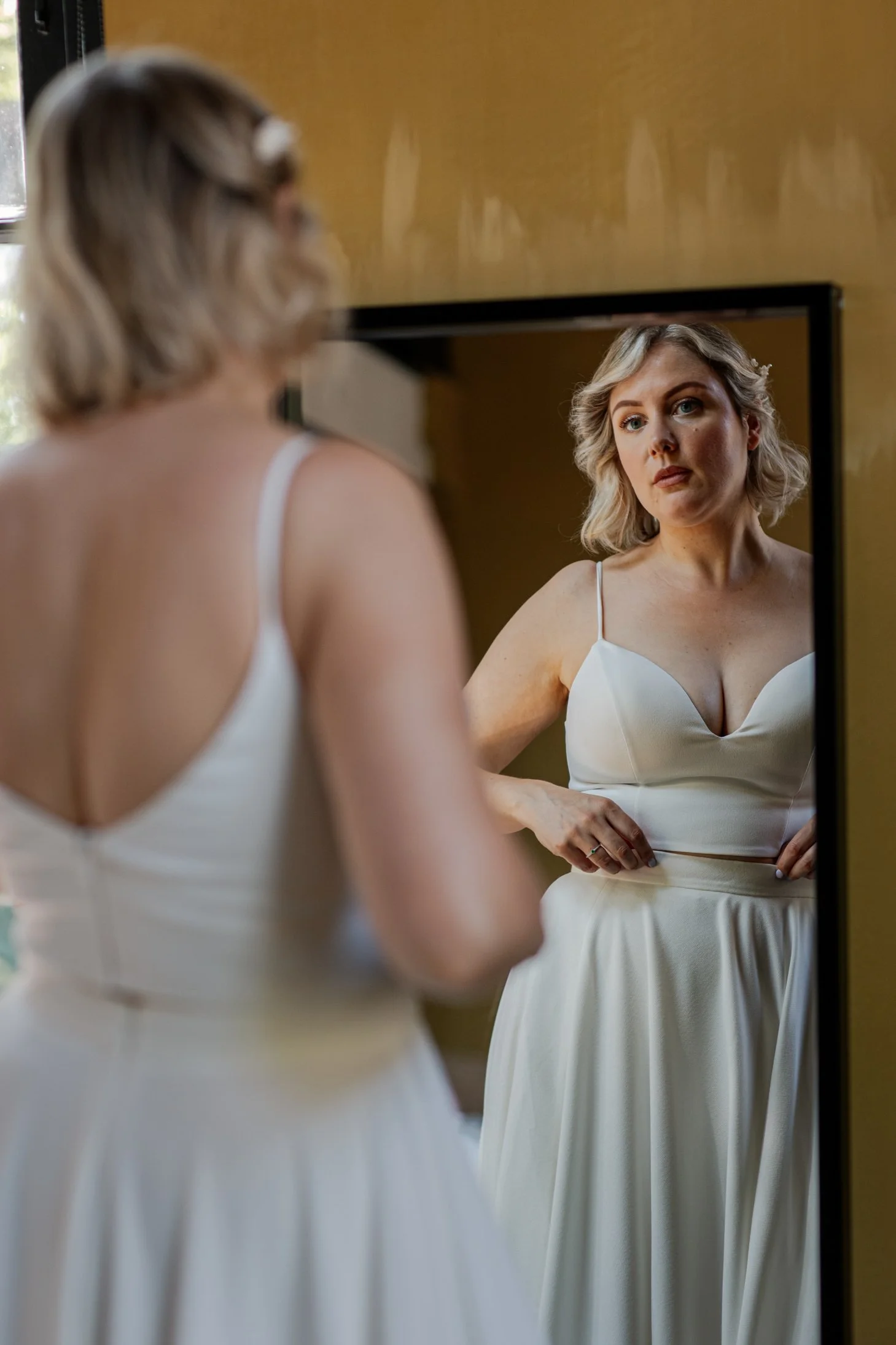 Een vrouw bekijkt zichzelf in een spiegel, gekleed in een witte jurk, met een Spring Vrouw in de achtergrond.