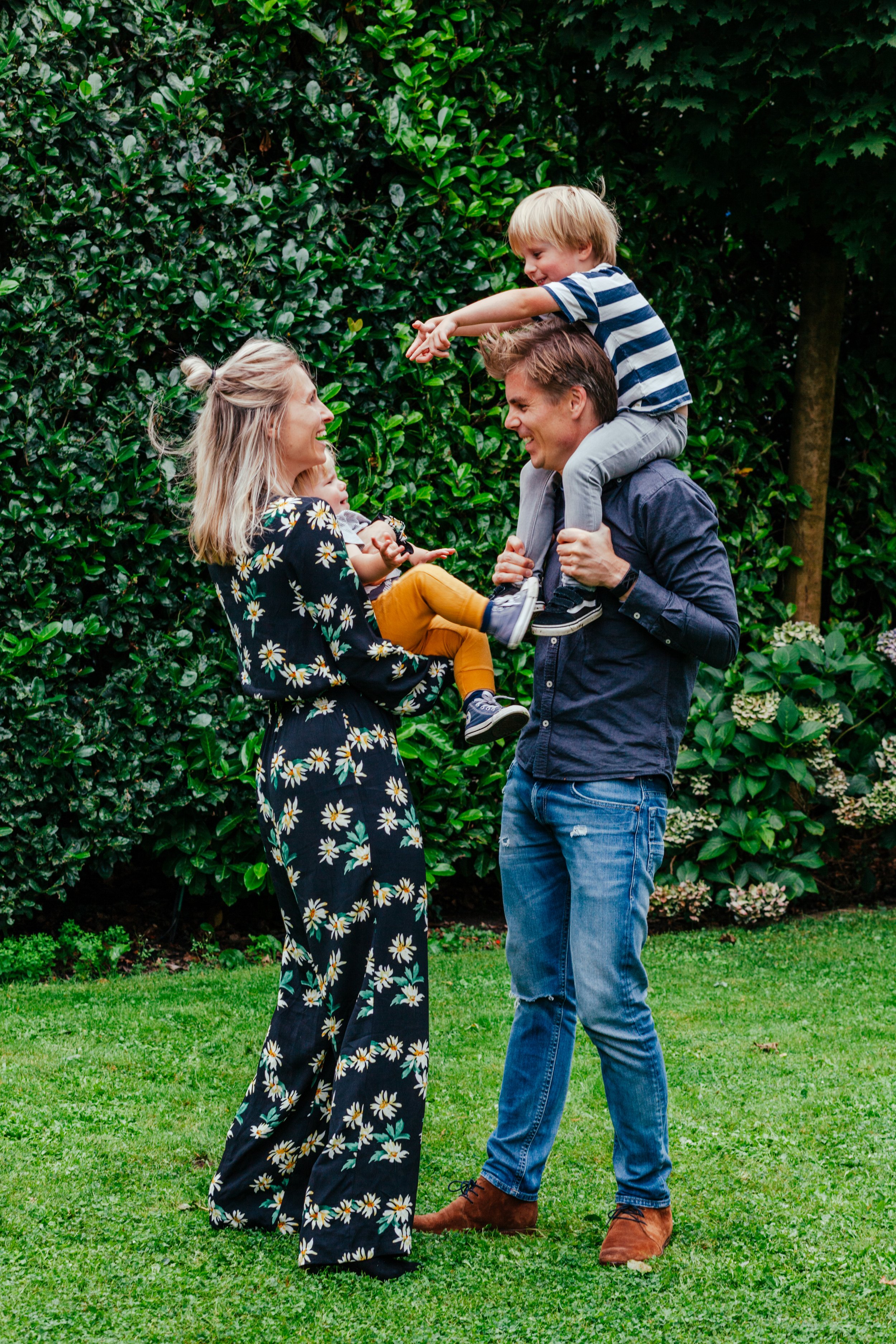 Een gelukkig gezinsmoment in een tuin met groene struiken en gras, waarbij een vrouw, man en twee jonge kinderen plezier maken.