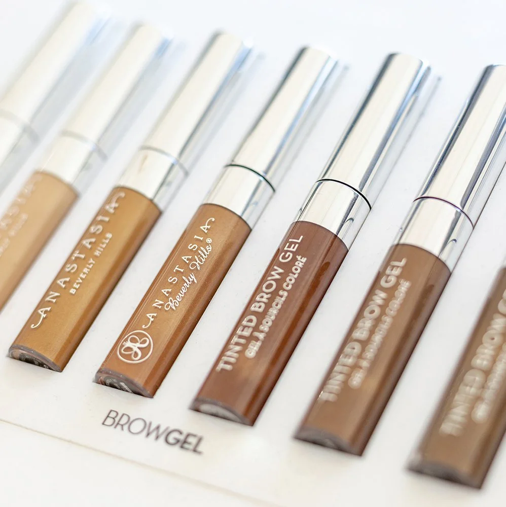 Verschillende kleuren wenkbrauwgel in doorzichtig buisje met zilveren dop, liggend op een wit oppervlak.