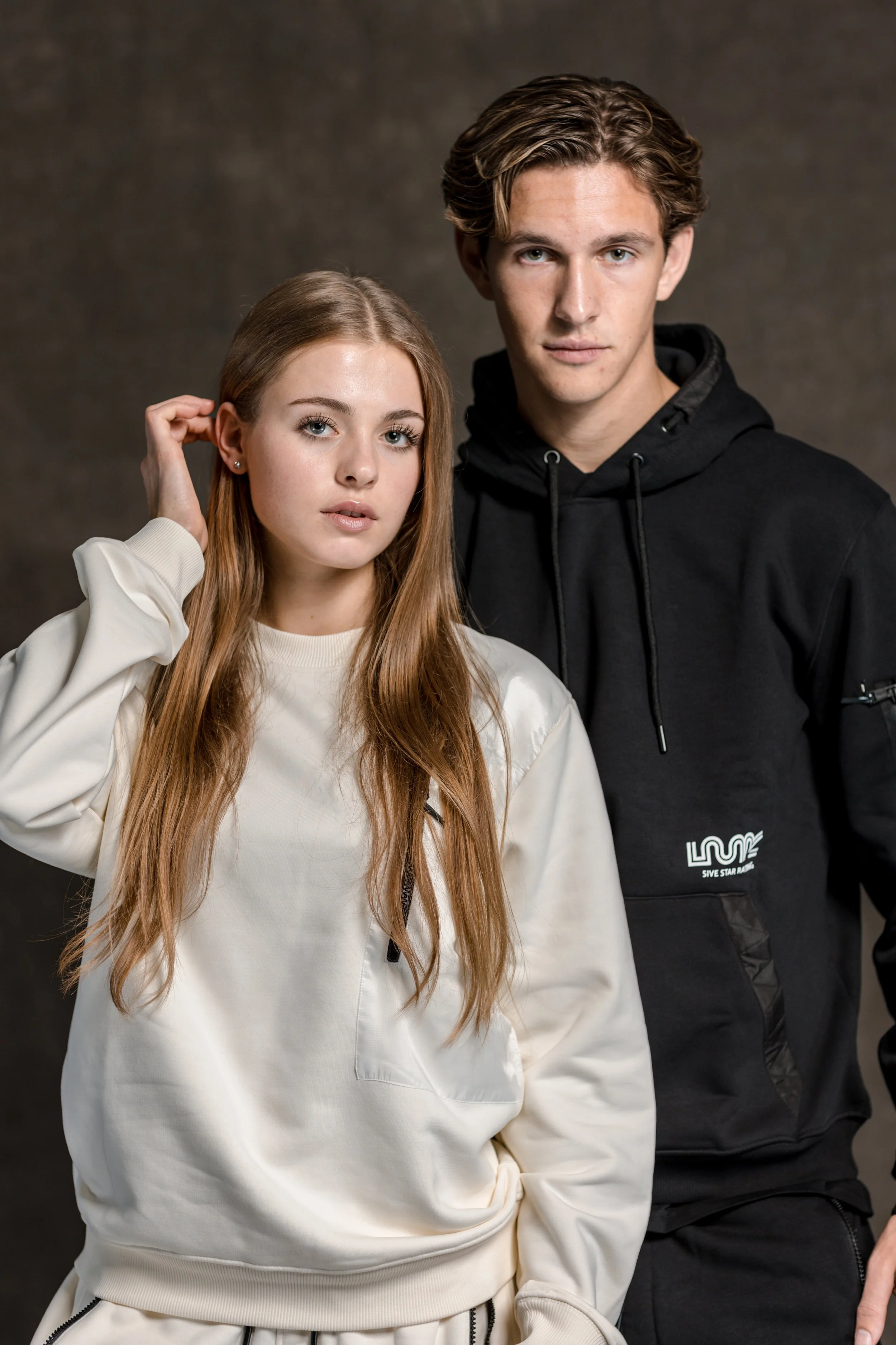 Twee jonge mensen, een vrouw en een man, poseert samen tegen een grijze achtergrond, beiden in casual sportkleding.
