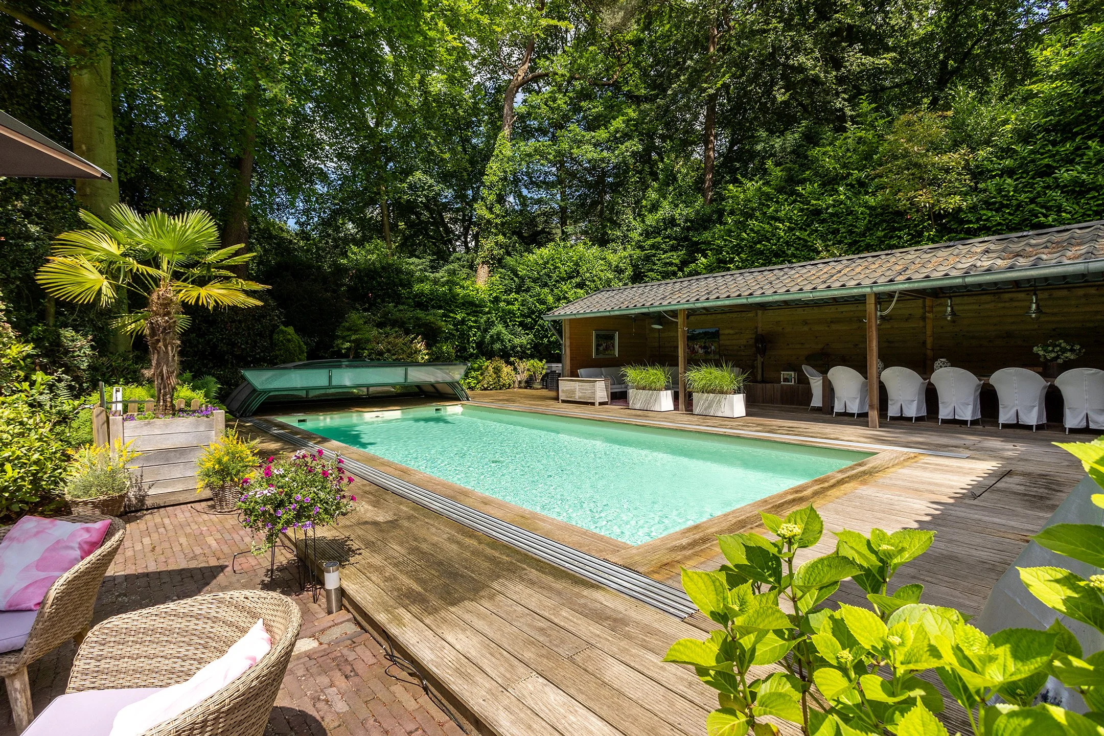 Luxe zwembad met houten dek, omgeven door groene bomen en planten, met een overdekte zithoek en een loungehoek met wicker stoelen en kussens.