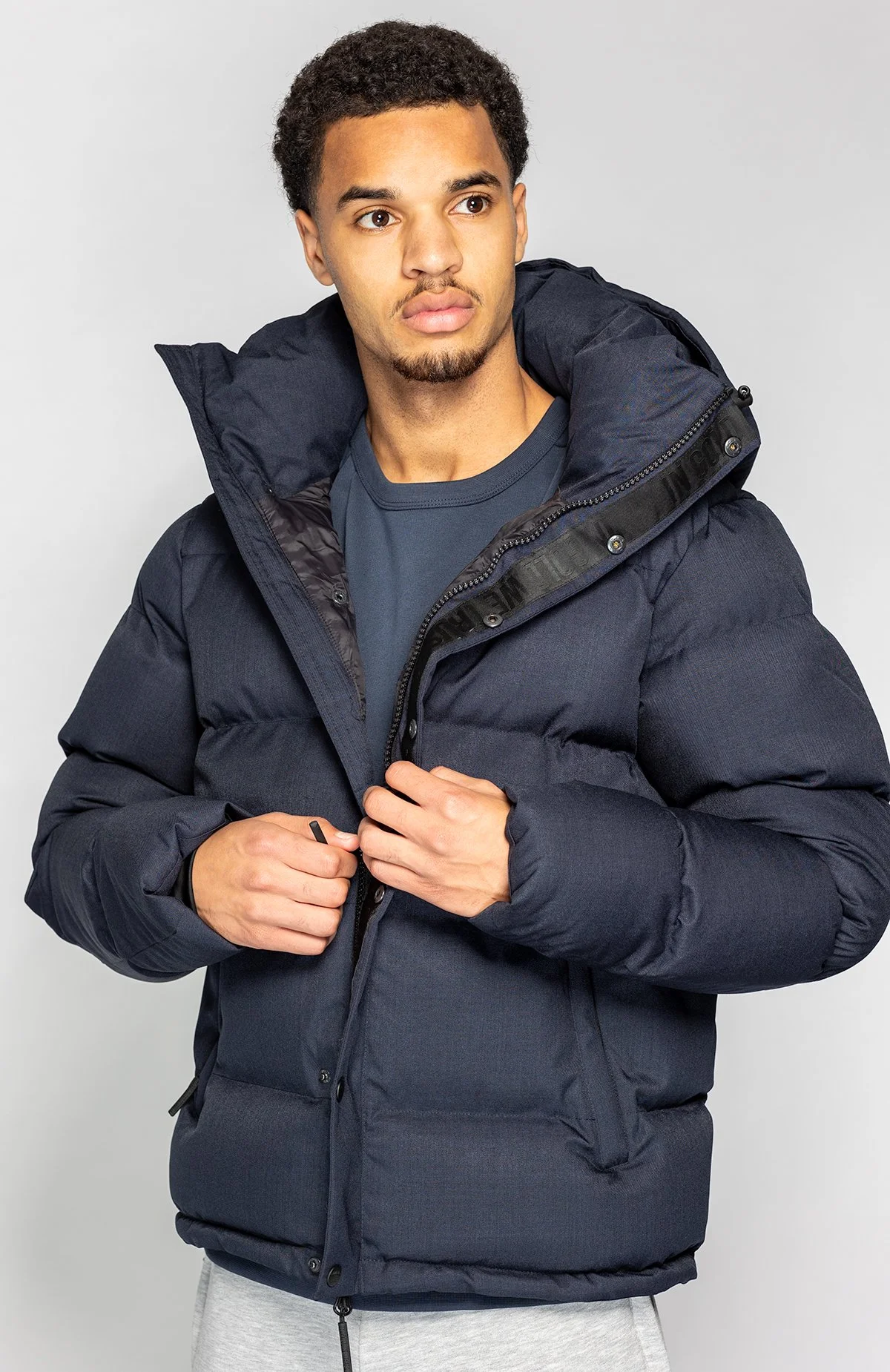 Man draagt een donkere, gewatteerde winterjas met capuchon, staand tegen een lichte achtergrond.