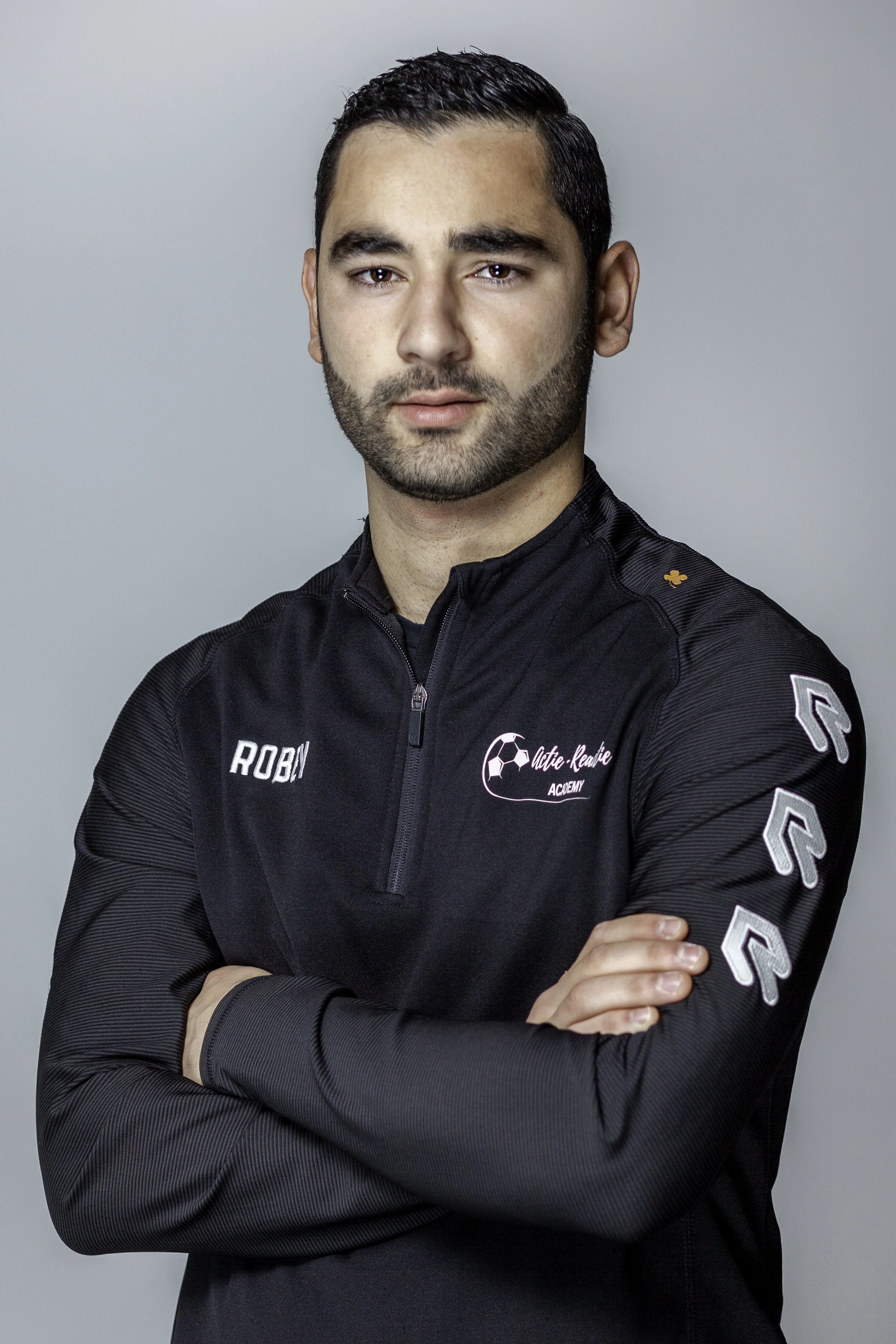 Een man met kort zwart haar en een getrimde baard, gekleed in een zwart sportjack met bedrijfslogo's en tekst, die tegen een lichte, effen achtergrond staat.