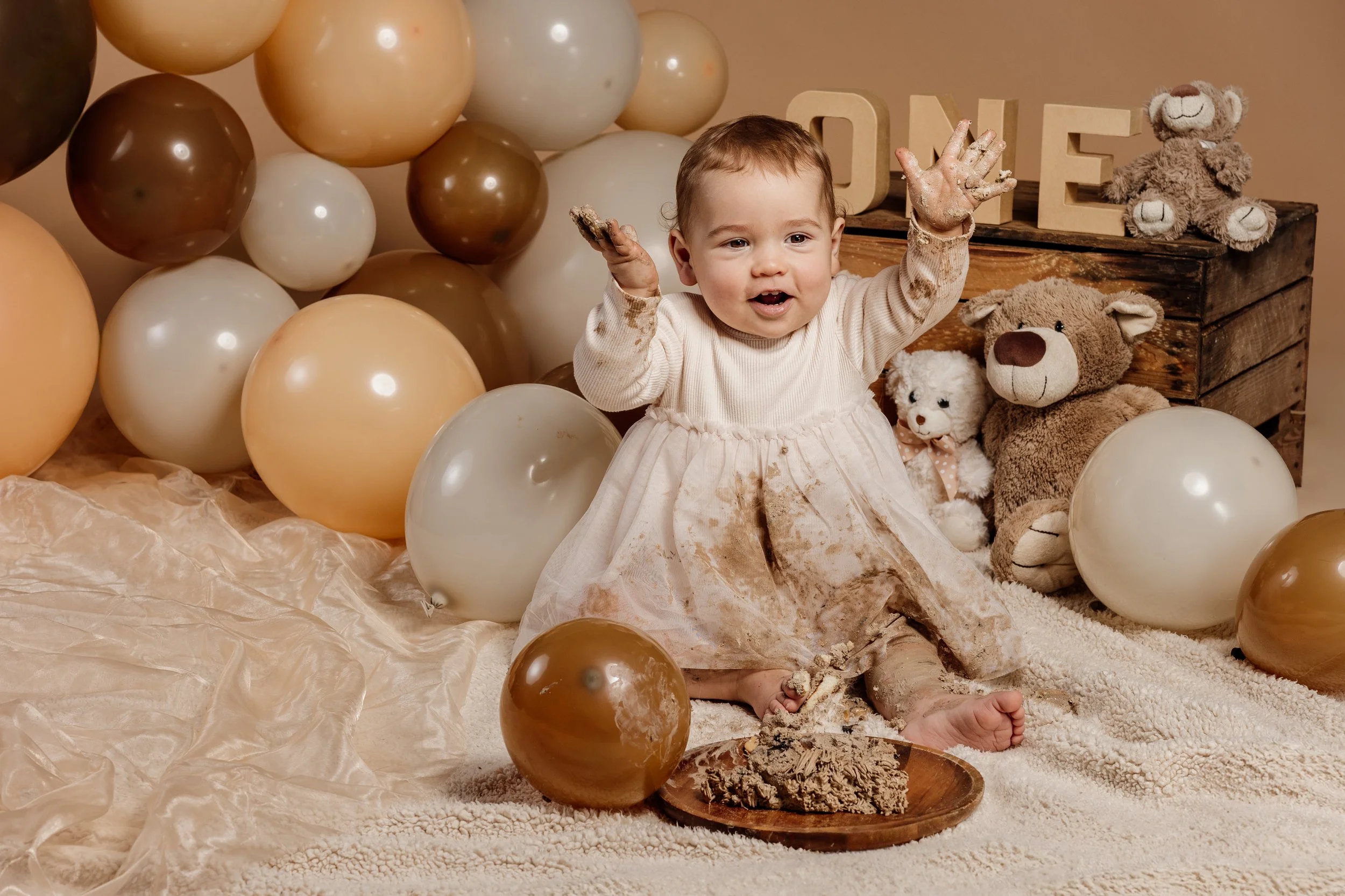 Lachend peuterknul die in een crèmekleurige jurk zit, omringd door ballonnen en teddyberen, met modder op haar handen en kleren, zittend op een zachte beige deken, met letterstenen en een houten kist op de achtergrond.