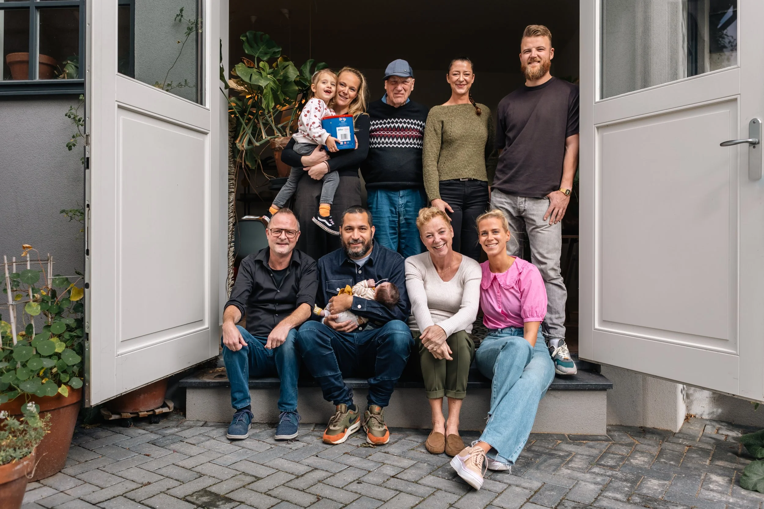 Een grote groep mensen, vermoedelijk familie, poseert voor de voordeur van een huis. Er zijn vier volwassenen en vier kinderen, waaronder een baby en een peuter. Ze lijken blij en ontspannen.