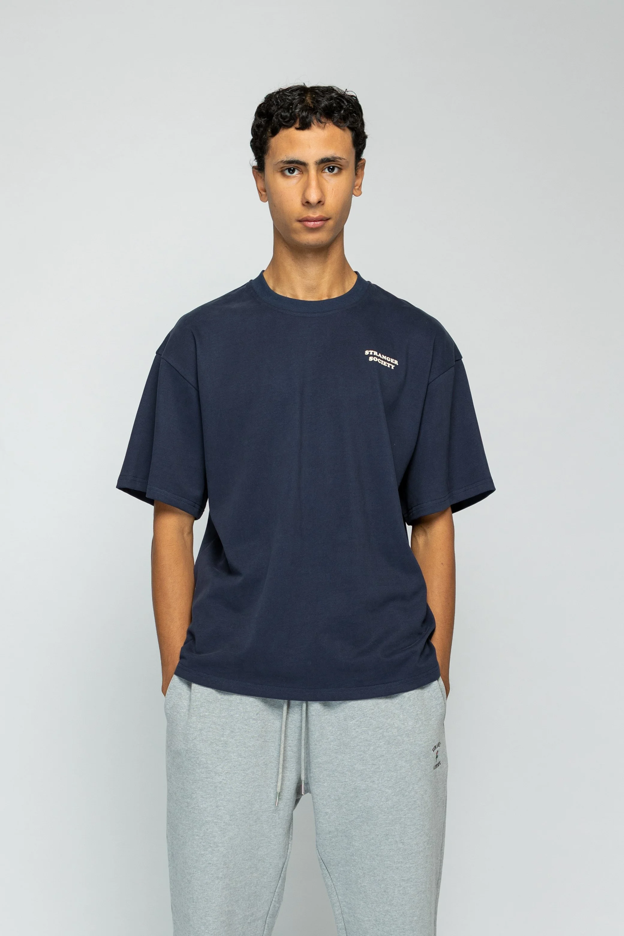 Jonge man in een donkere T-shirt en grijze sweatpants staat rechtop tegen een witte achtergrond.