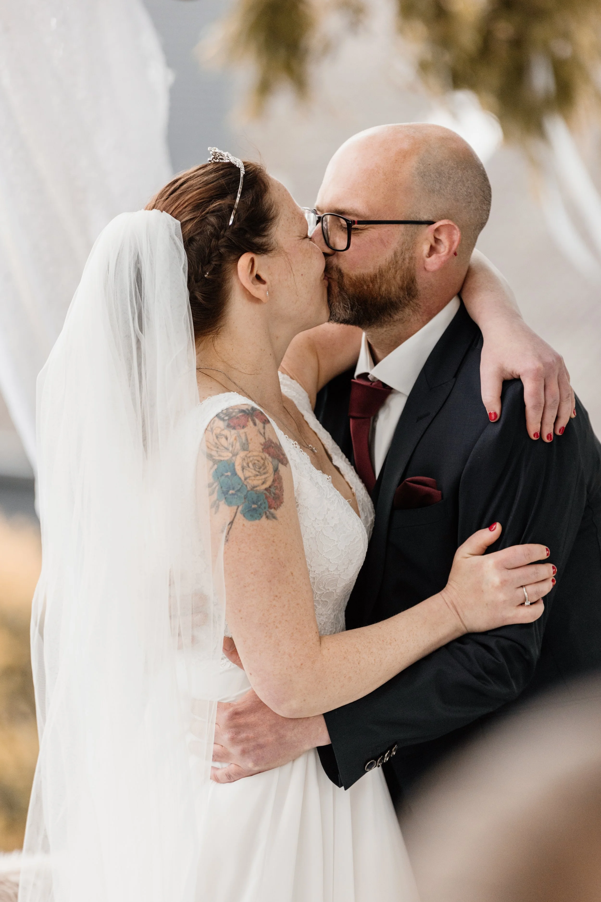 Bruidegom en bruid kussen elkaar tijdens hun huwelijk, zij draagt een witte jurk met tatoeages op haar arm, hij draagt een zwart pak en bril.