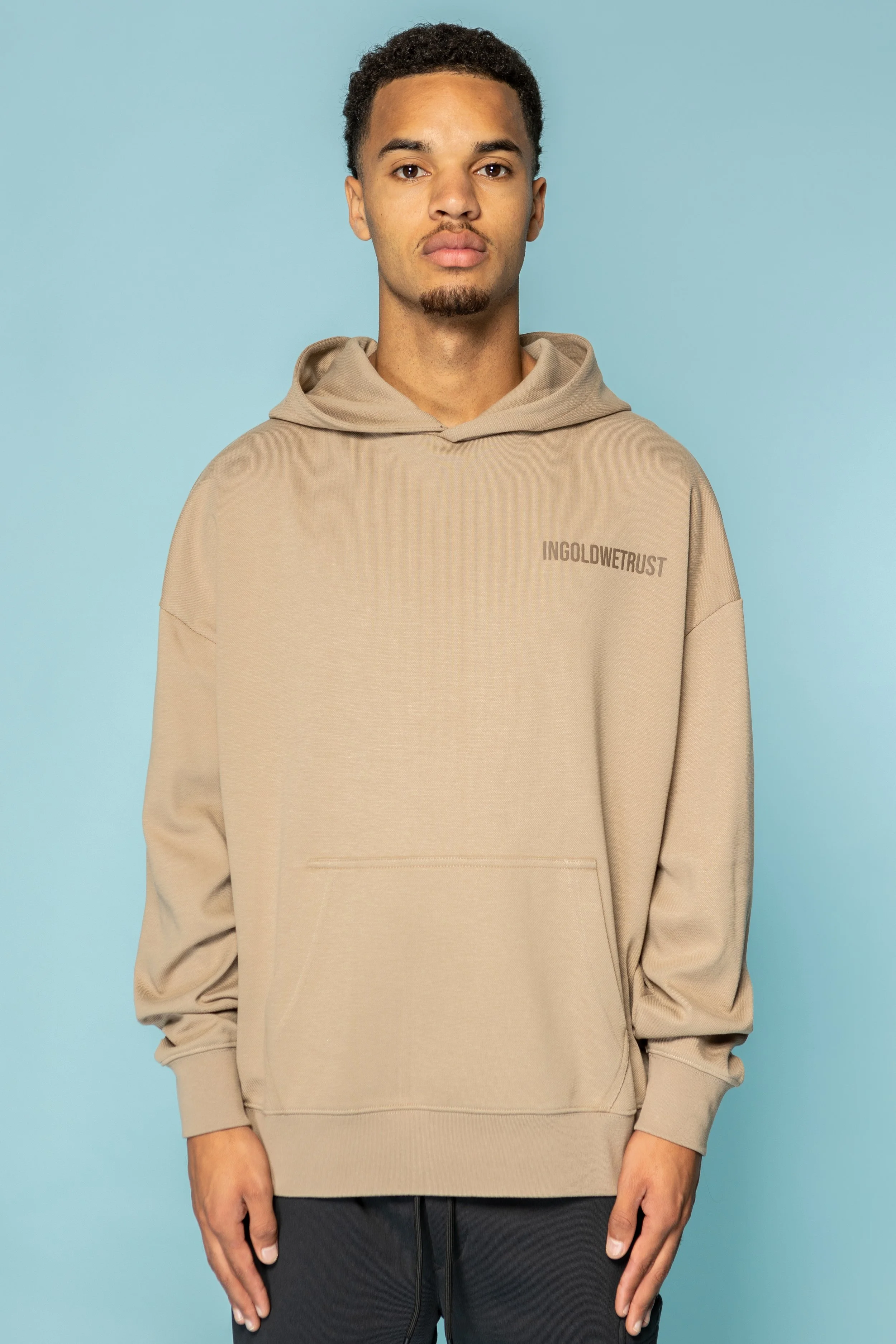 Man draagt een beige hoodie met de tekst 'INGOLDWTRUST' op de linkerbovenkant. Onderaan een ruime zak, tegen een lichtblauwe achtergrond.