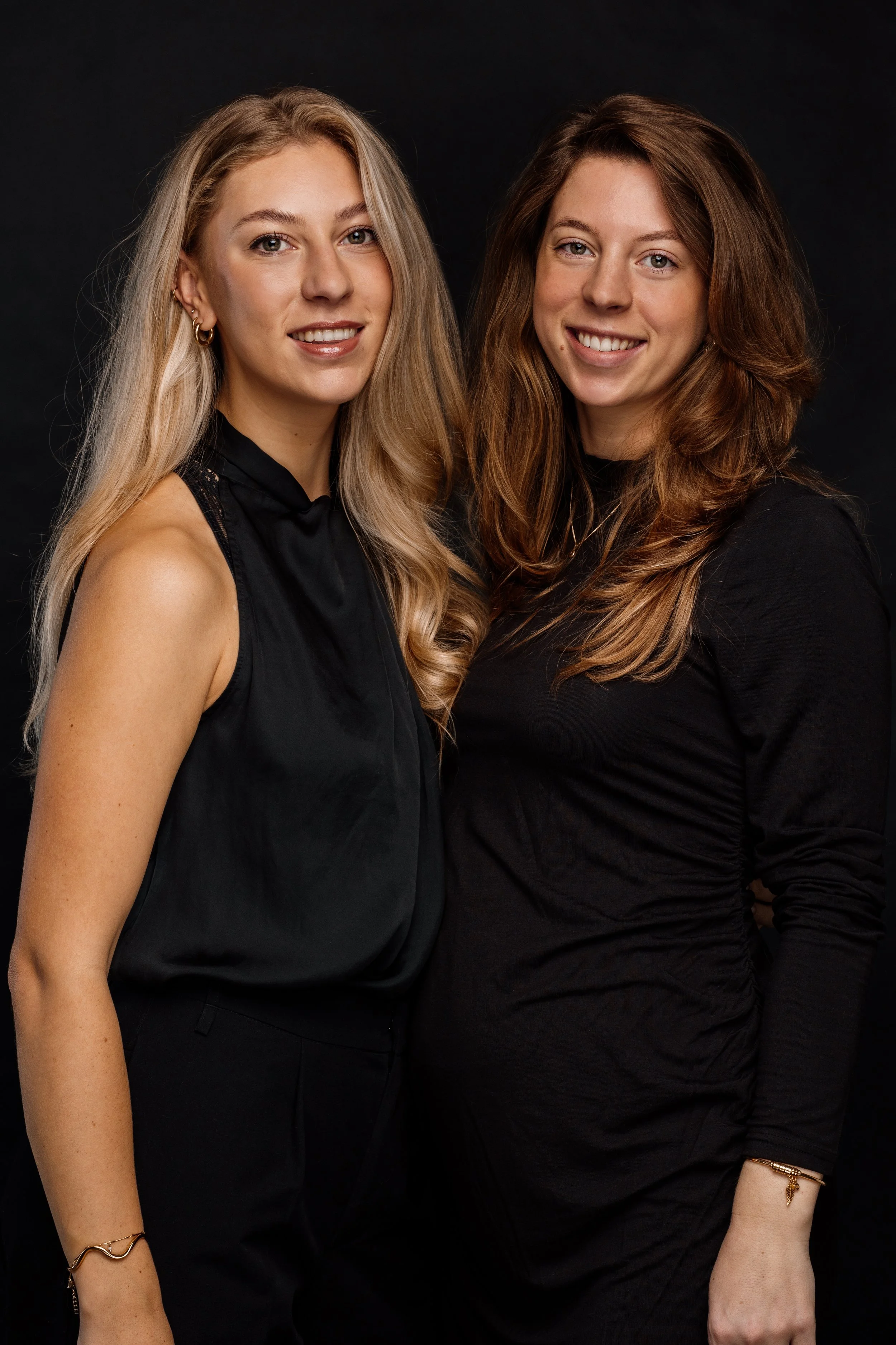 Twee vrouwen met lang haar, glimlachend, staand dicht bij elkaar, gekleed in zwart, tegen een zwarte achtergrond.