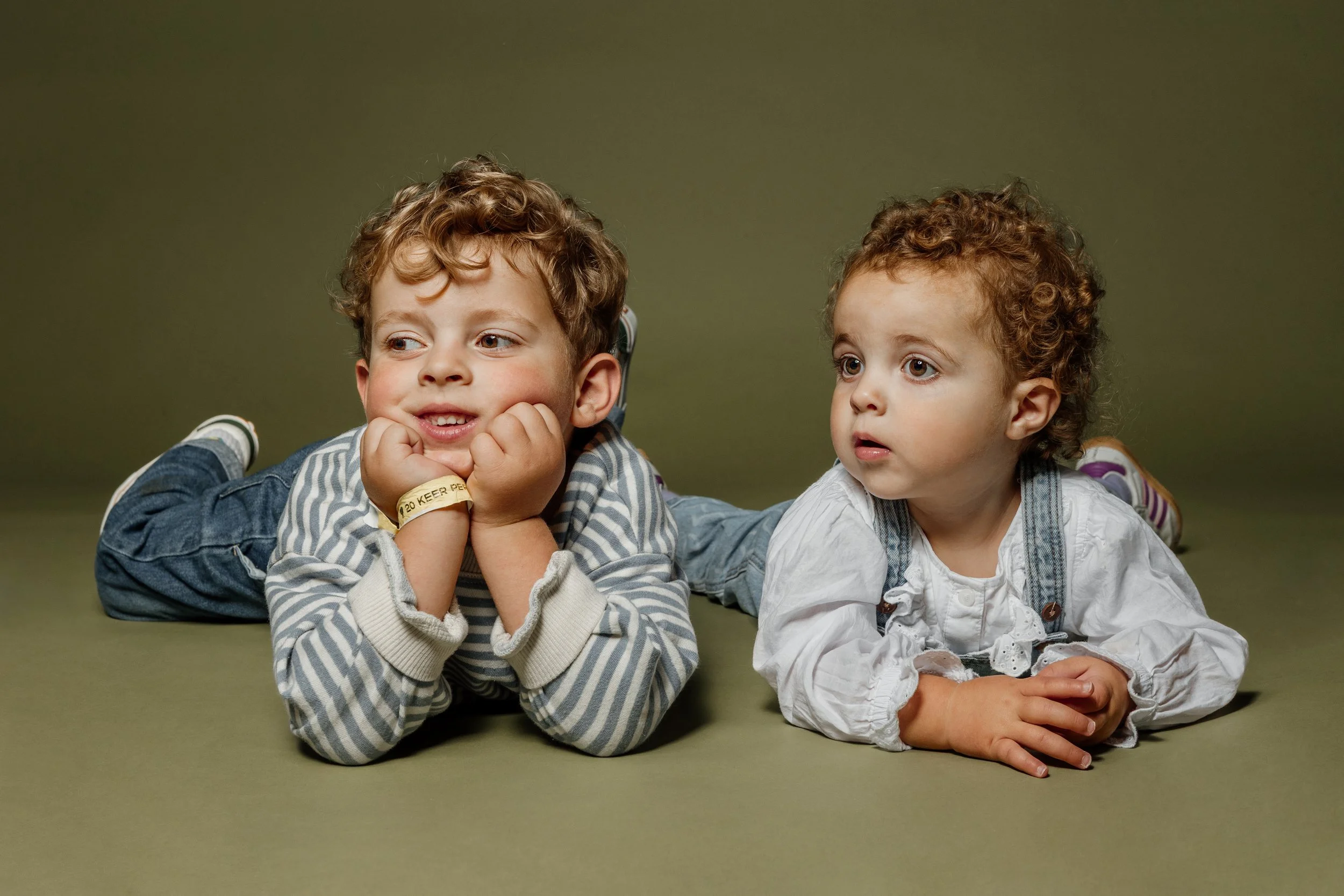 Twee jonge kinderen liggen op de grond, één met hoofd in handen, de ander kijkt nieuwsgierig.