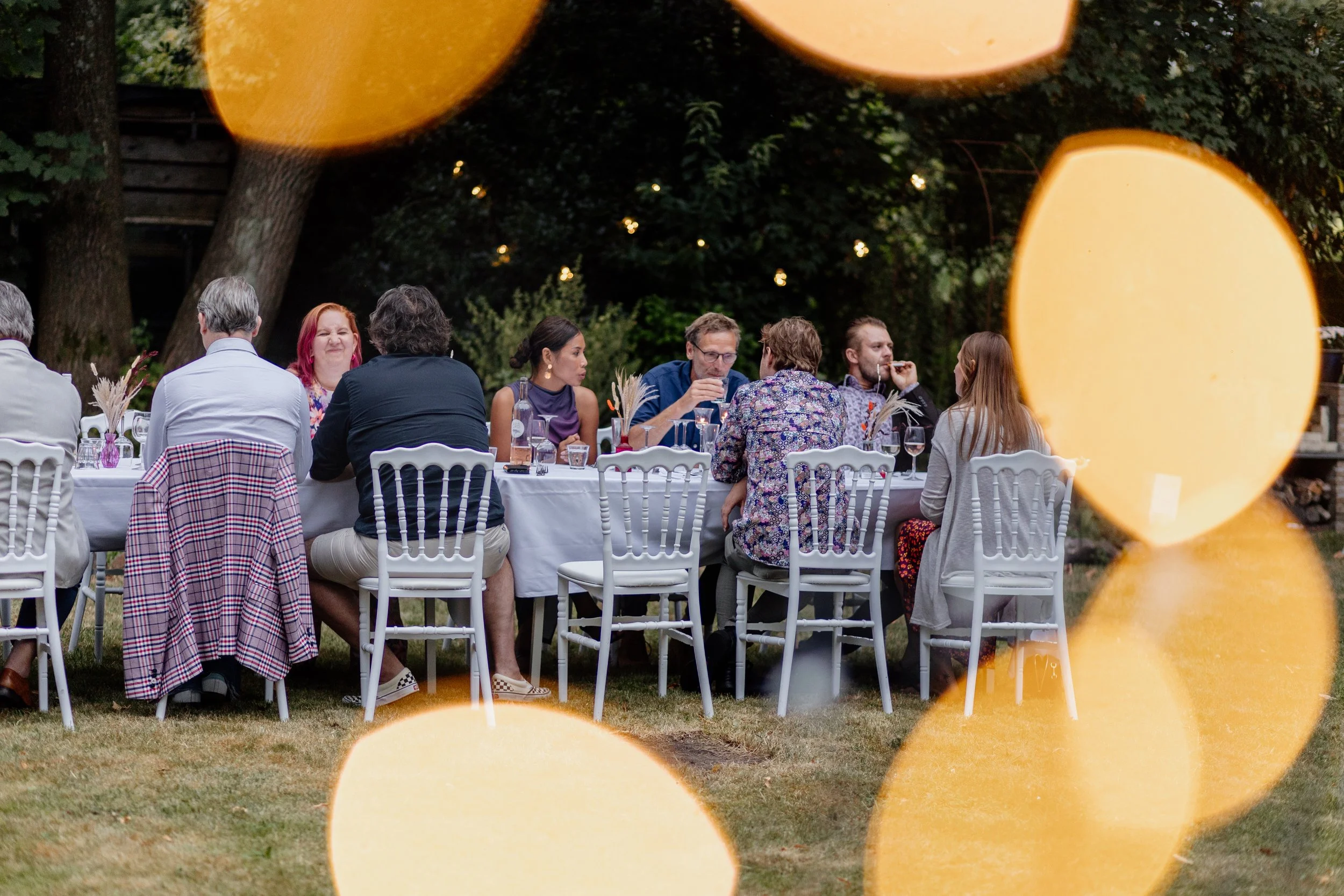Gasten zitten aan een lange tafel met witte borden en glazen tijdens een buitenevenement 's avonds, met bomen op de achtergrond.
