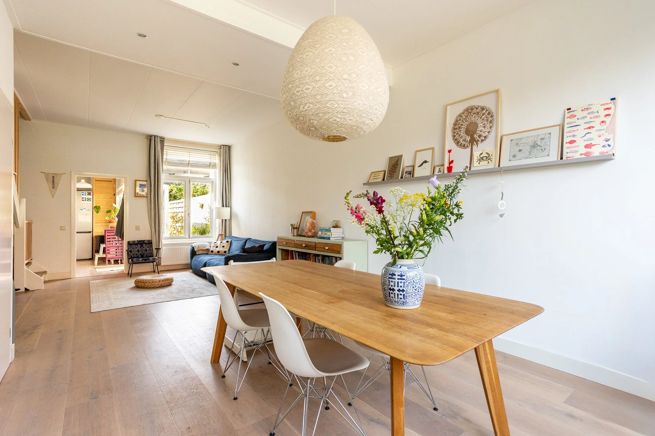 Interieur van een woonkamer met een eettafel, bloemenvaas, bank, muurdecoraties en een grote ramen met uitzicht op de tuin.
