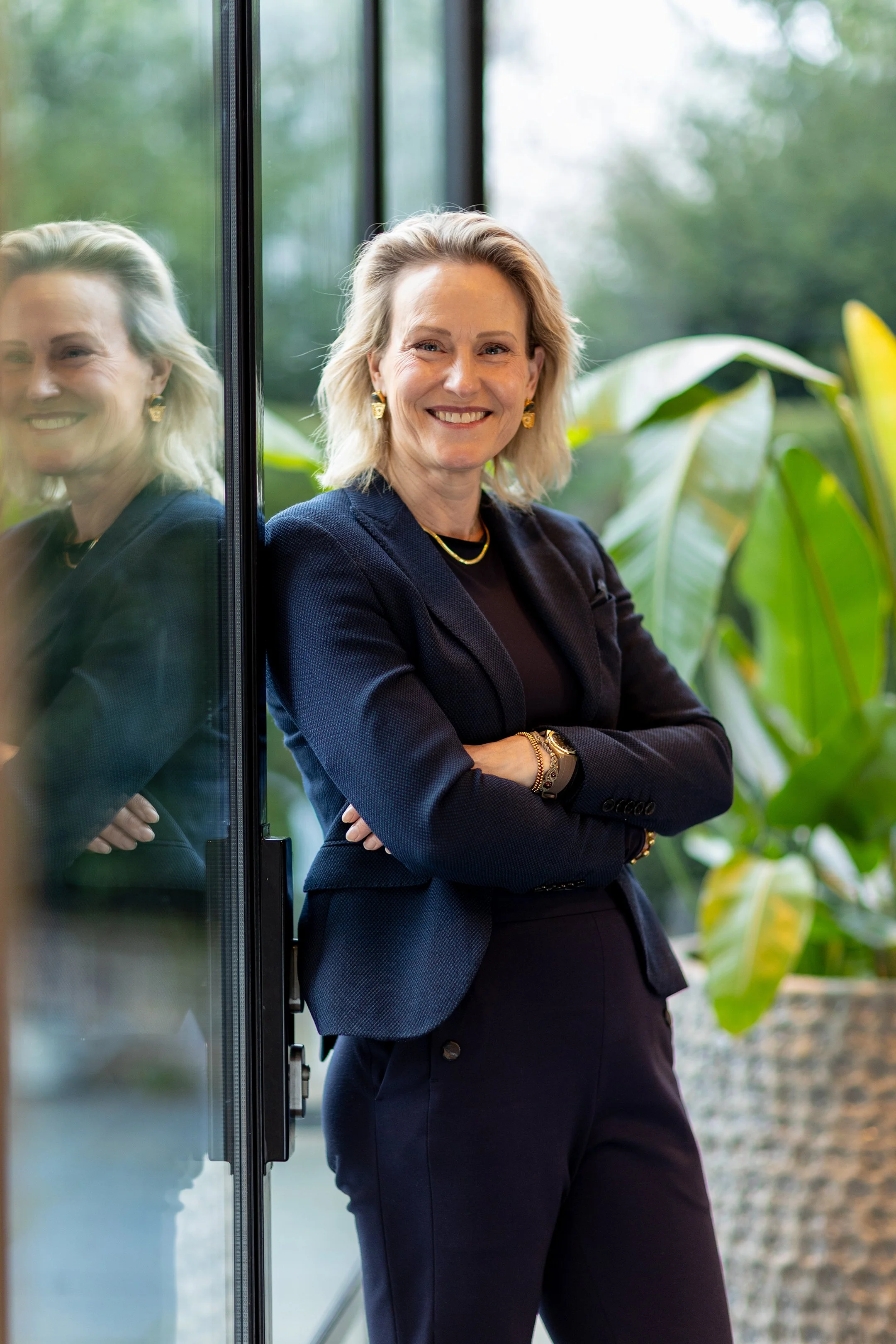 Een lachende vrouw in een zakelijke outfit staat met armen over elkaar naast een glazen deur, met groen planten en een bosrijke achtergrond buiten.