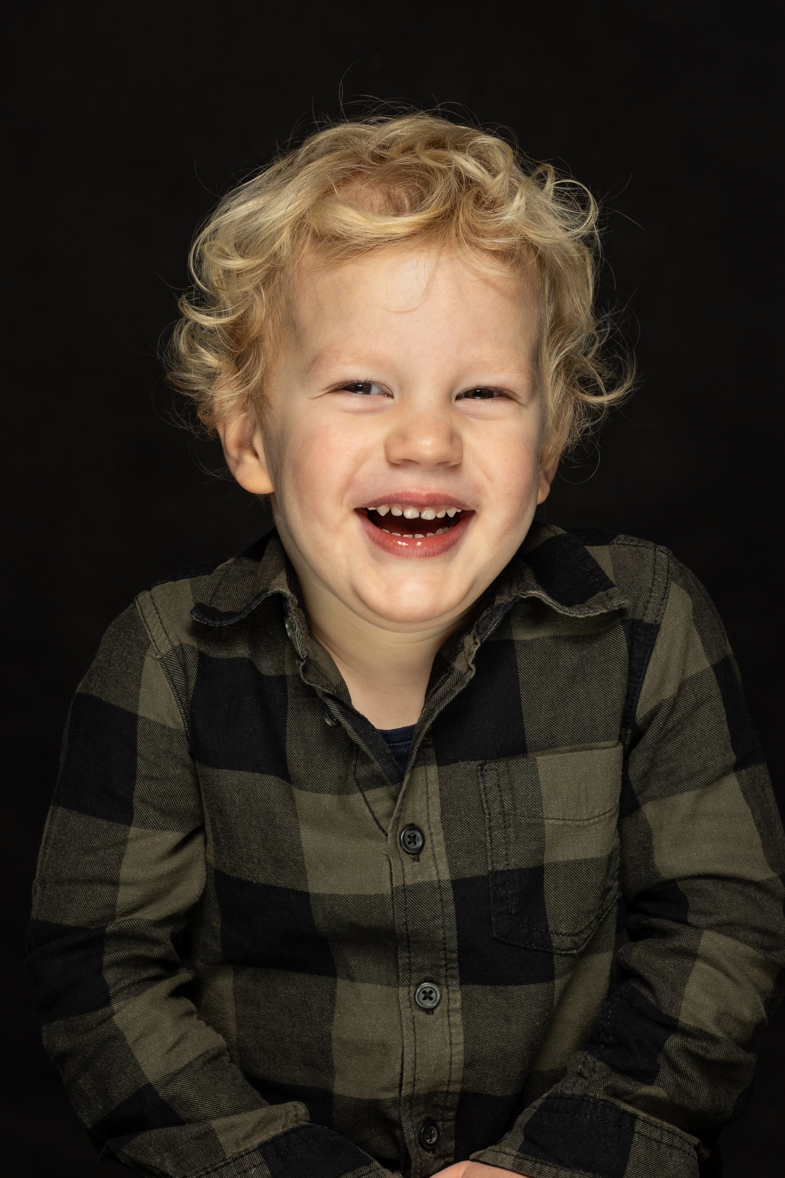 Een lachende jonge jongen met blond, krullend haar draagt een groen en zwart ruitjes overhemd, tegen een zwarte achtergrond.