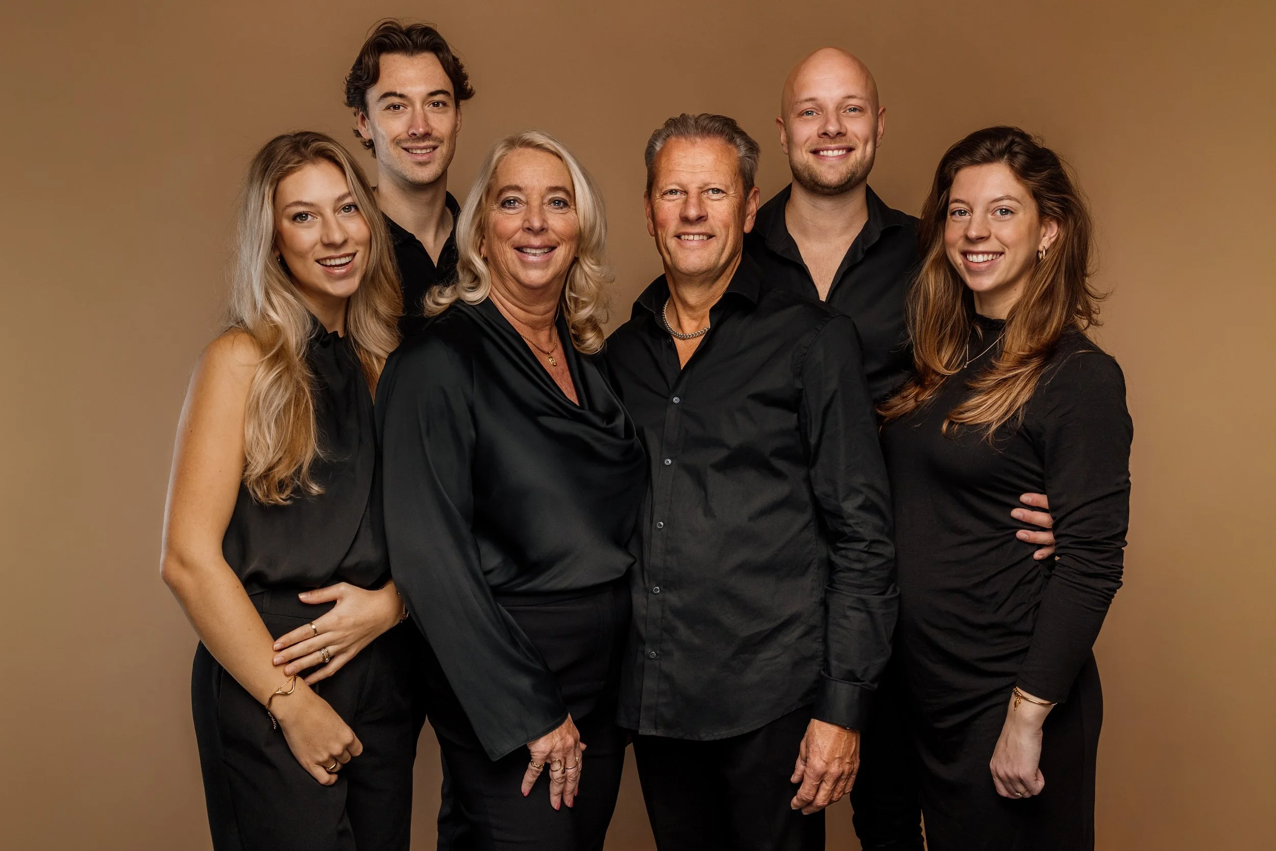 Zes mensen, drie mannen en drie vrouwen, poseerden samen voor een kamer. Ze dragen zwarte kleding tegen een beige achtergrond.