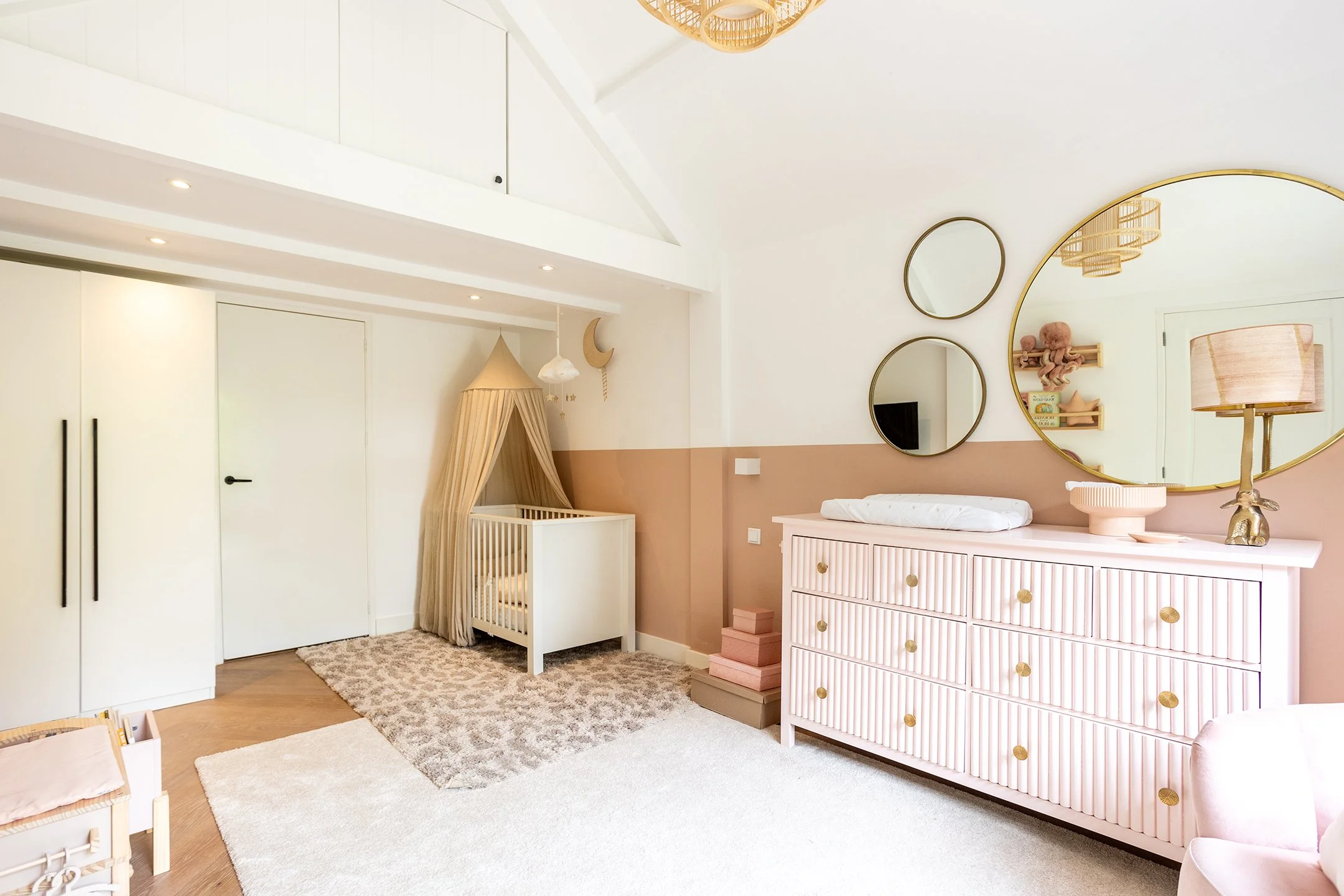 Een babykamer met pastelkleuren, een commode met gouden knoppen, een wieg met een gordijn, en decoratieve spiegels aan de muur.