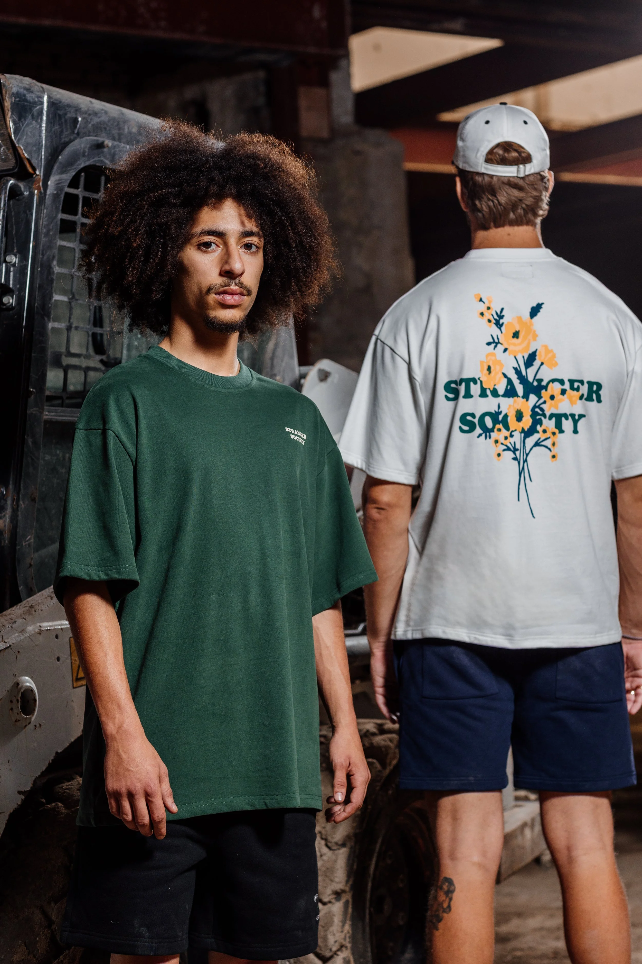 Twee jonge mannen staan in een industriële ruimte. Eén kijkt naar de camera, de ander heeft de rug naar de camera en draagt een witte T-shirt met een bloemenprint en de tekst 'STRONGER SOCIETY.' De man die naar de camera kijkt heeft een groene T-shir