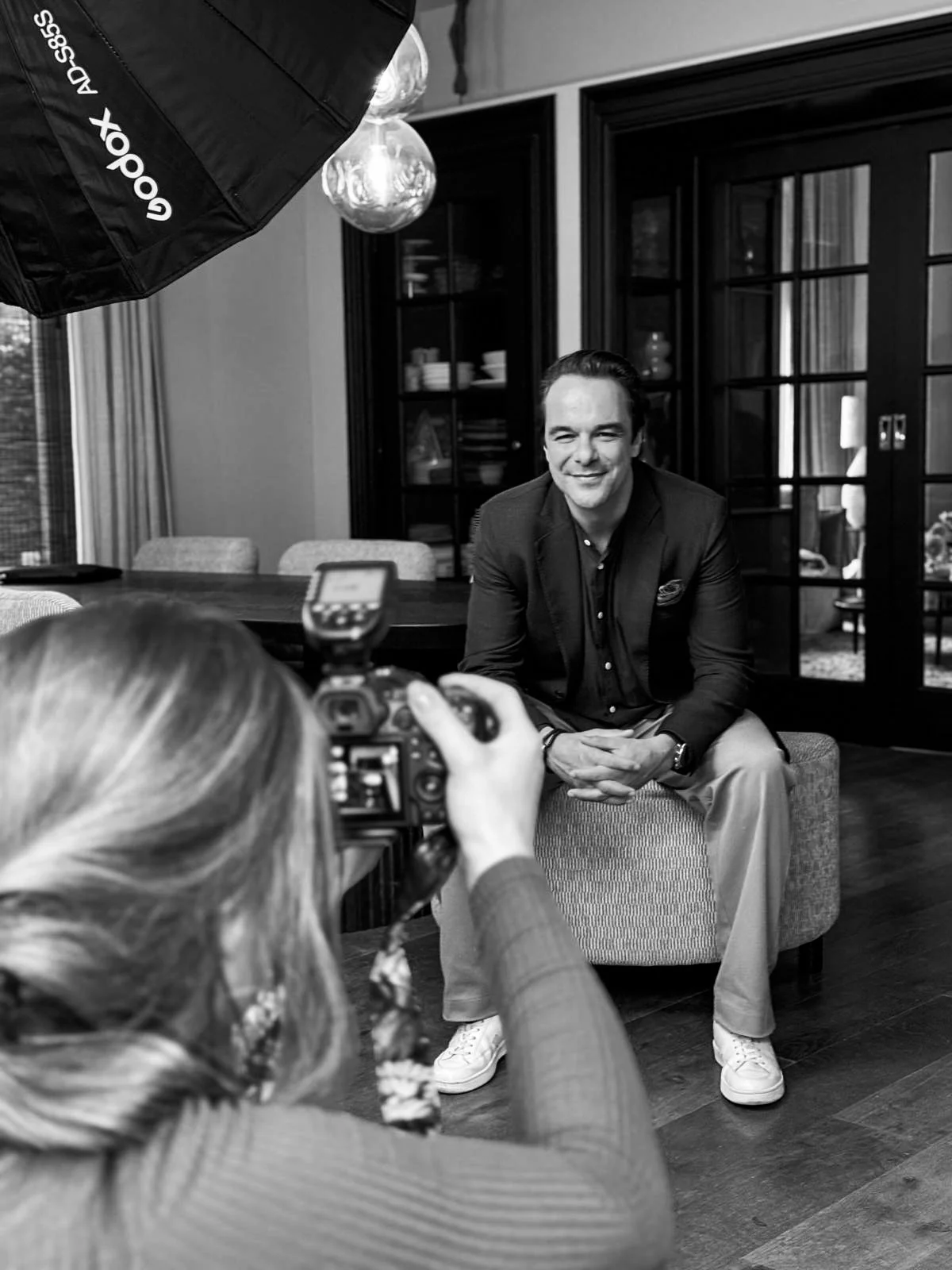 Man zit op een stoel terwijl hij wordt gefotografeerd door een vrouw met een camera. Er is professionele verlichting zichtbaar.