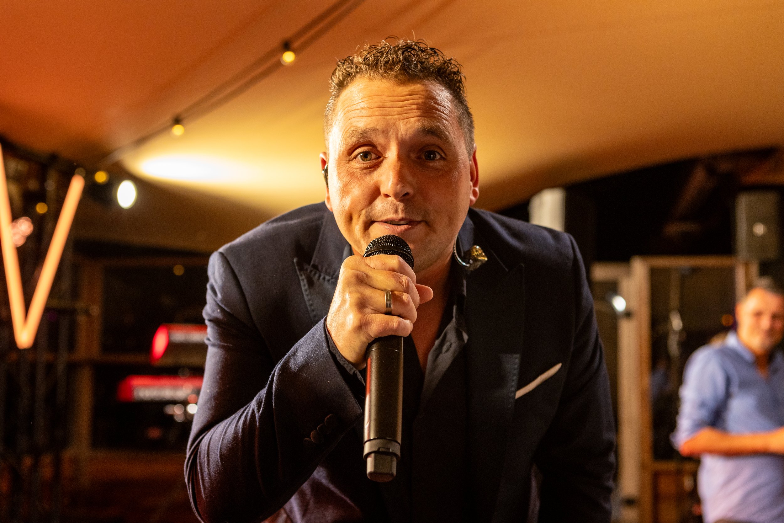 Man houdt microfoon vast en kijkt naar camera, op achtergrond een tweede man en muziekapparatuur, binnenshuis met warme verlichting.