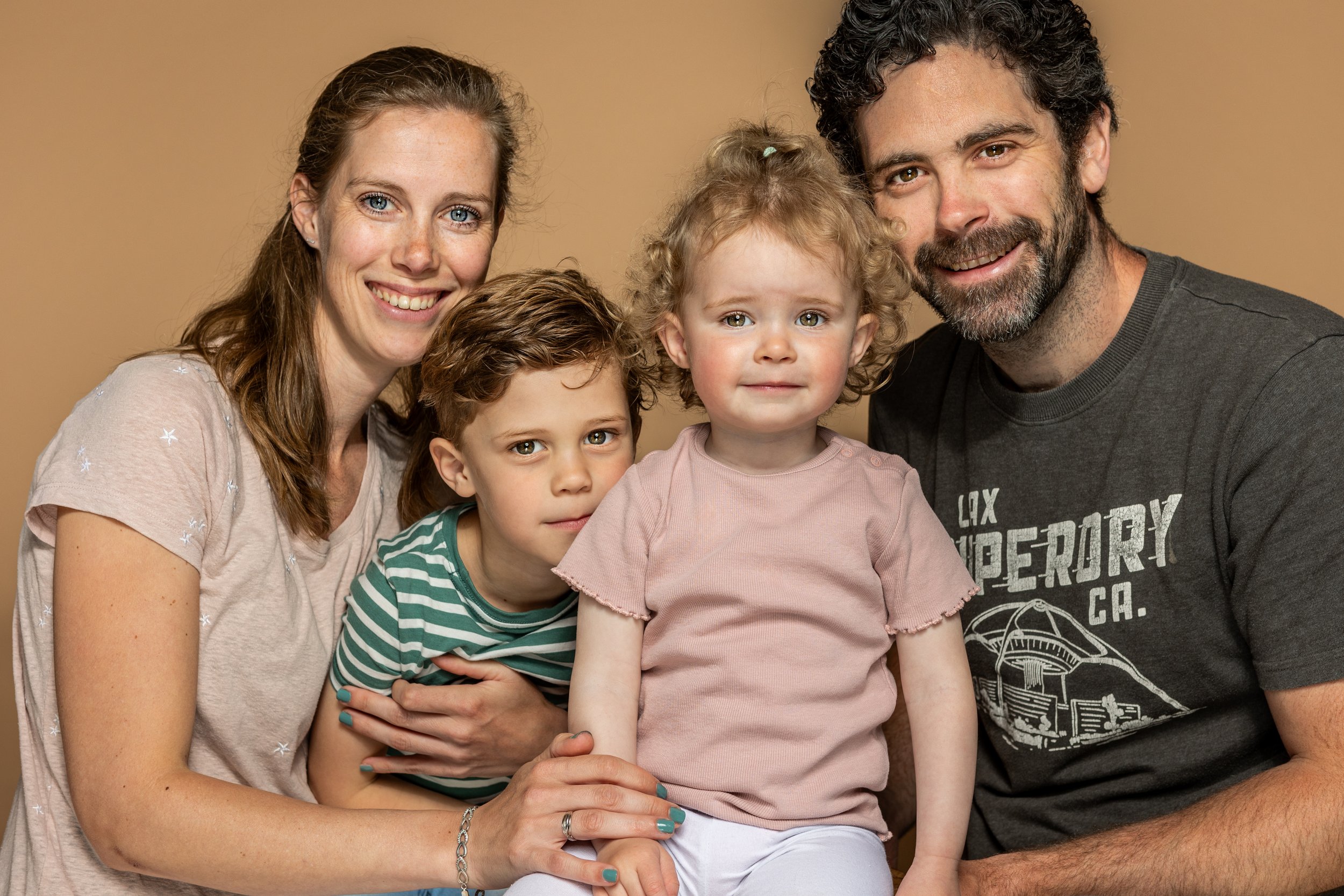 Familie van vier poseert samen tegen een beige achtergrond, lachend en knuffelend.