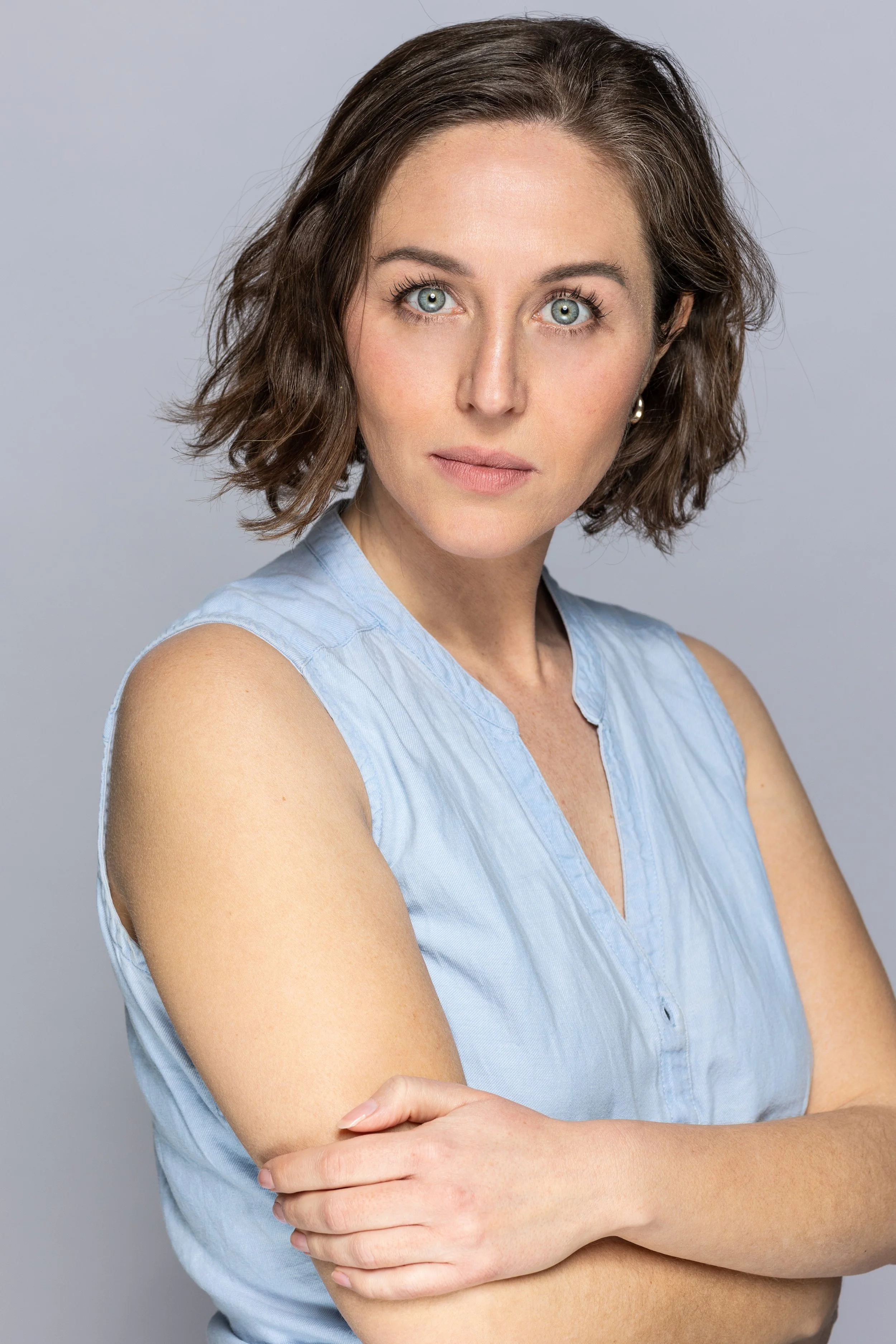 Portret van een vrouw met blauwe ogen en korte, golvende bruin haar, gekleed in een lichtblauw top, met een neutrale achtergrond.