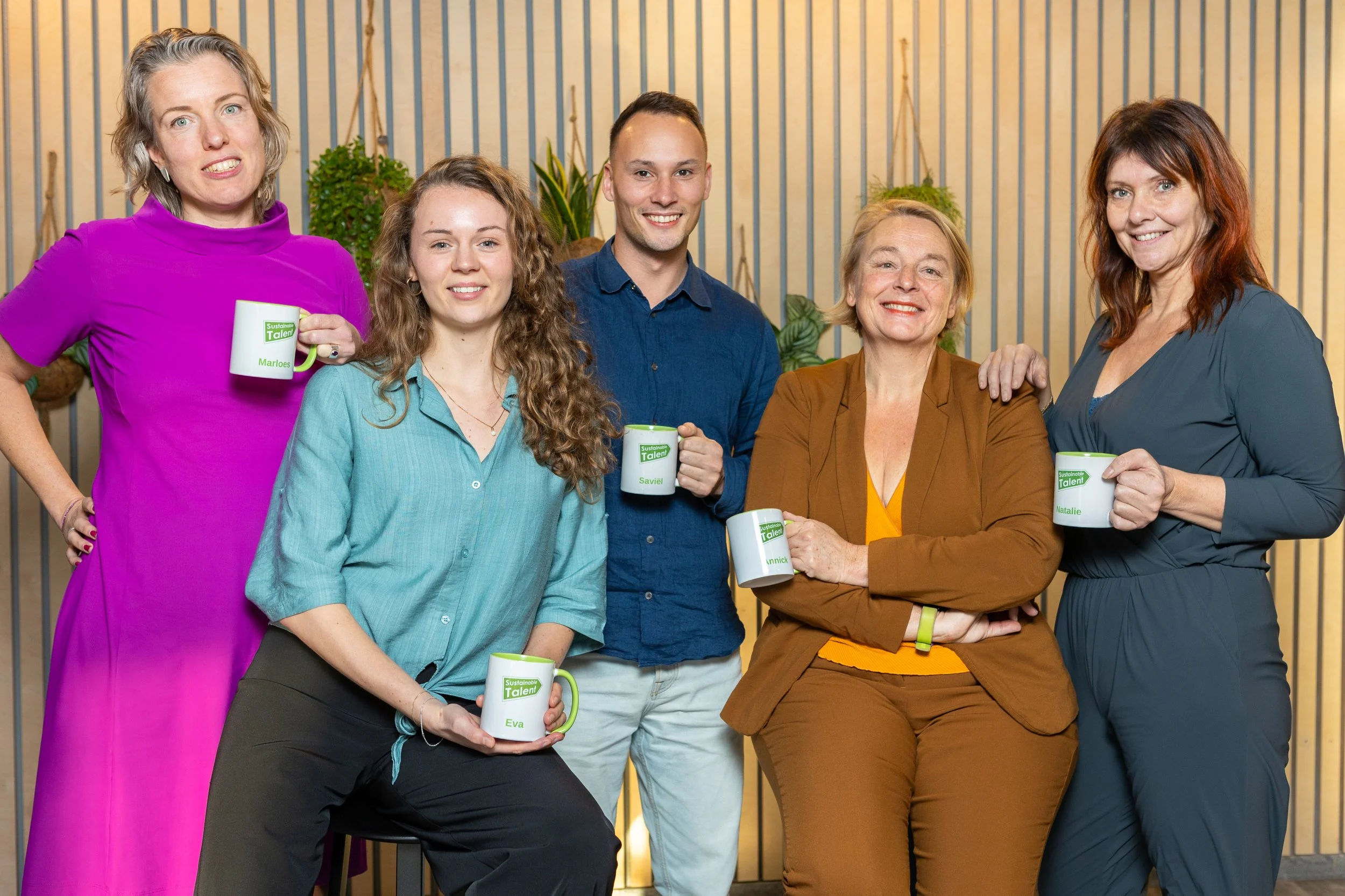 Zes mensen poseren samen in een interieur met planten op de achtergrond, en houden koffiemokken met het label 'Sustainable Talent' en namen erop.
