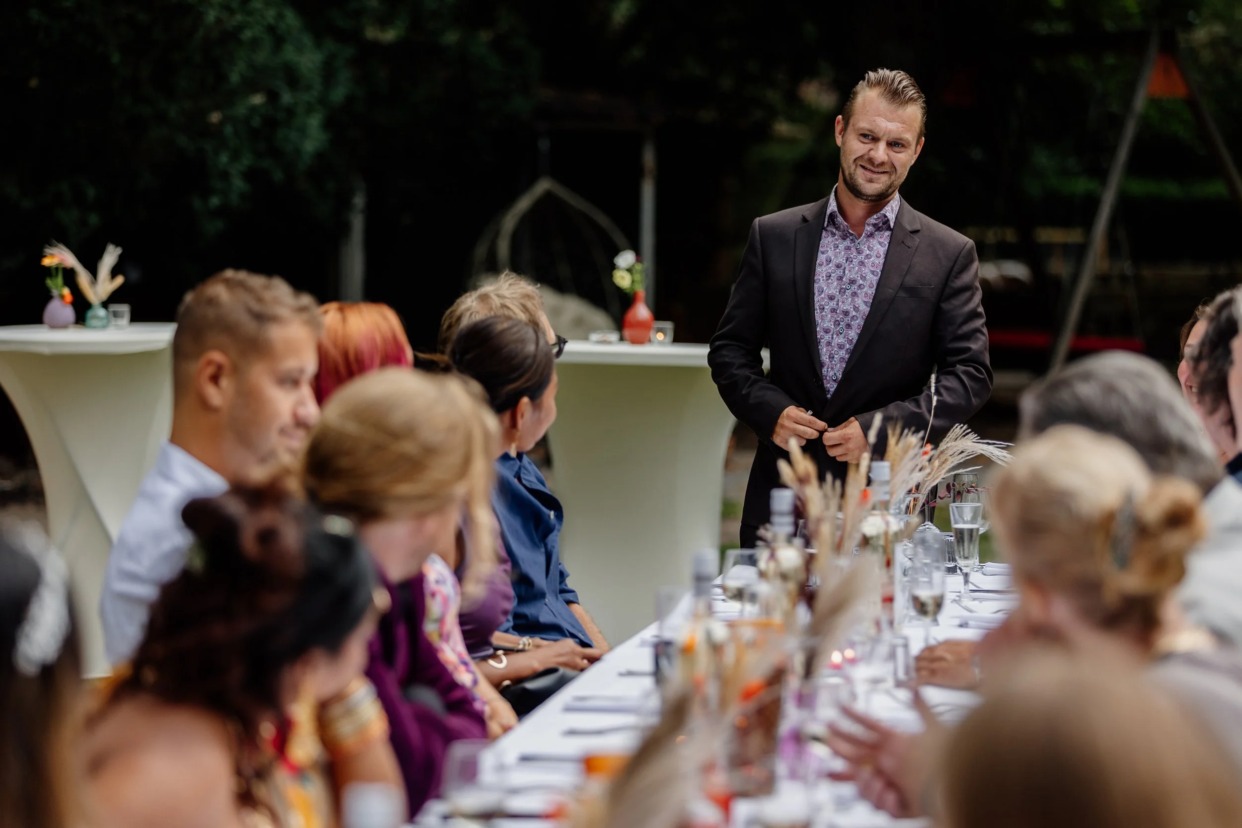 Man in donker pak geeft speech aan een lange tafel met gasten tijdens een buitengelegen evenement.