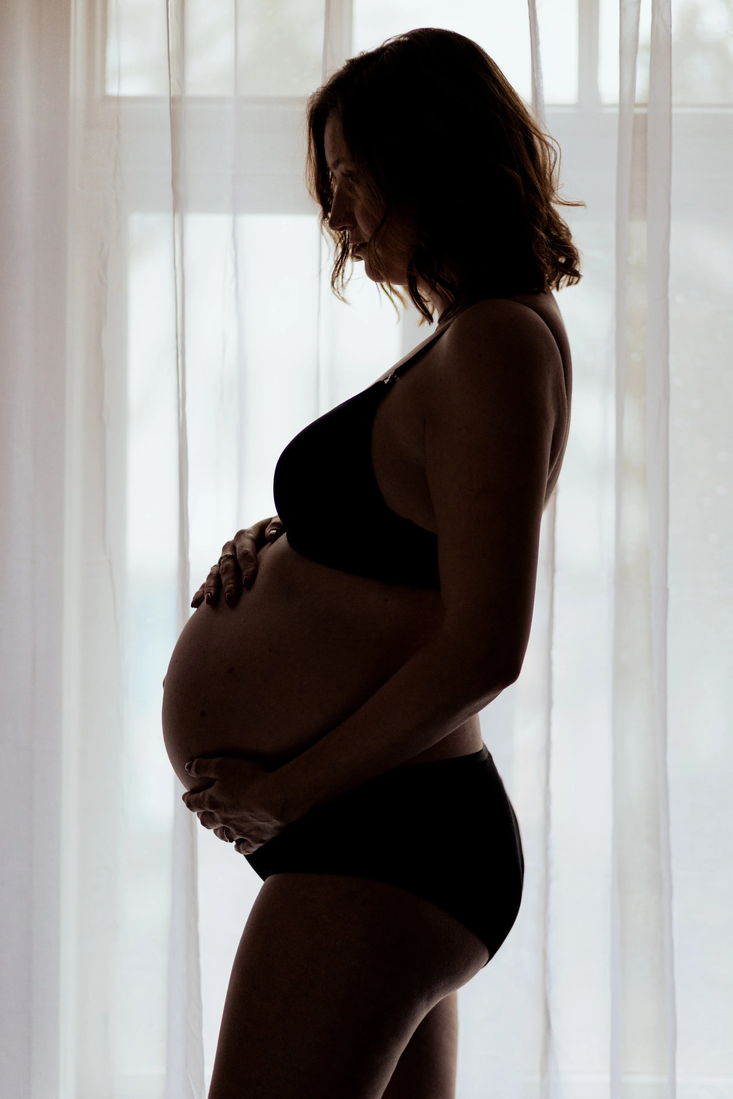 Silhouet van een zwangere vrouw die naar haar buik kijkt, staand voor een raam met witte gordijnen.