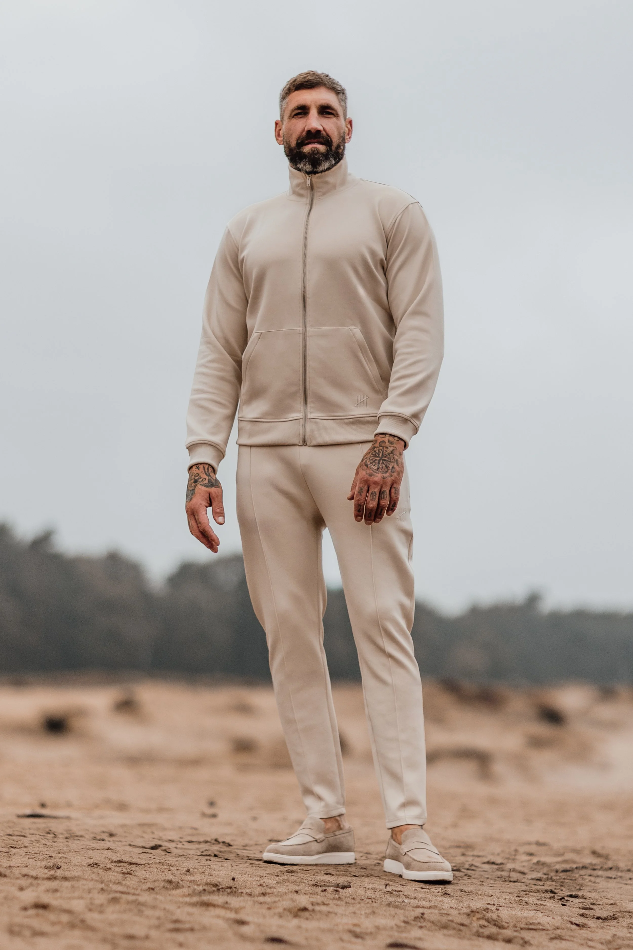 Man staat op een strand in een beige trainingspak en sneakers, met een bewolkte hemel op de achtergrond.