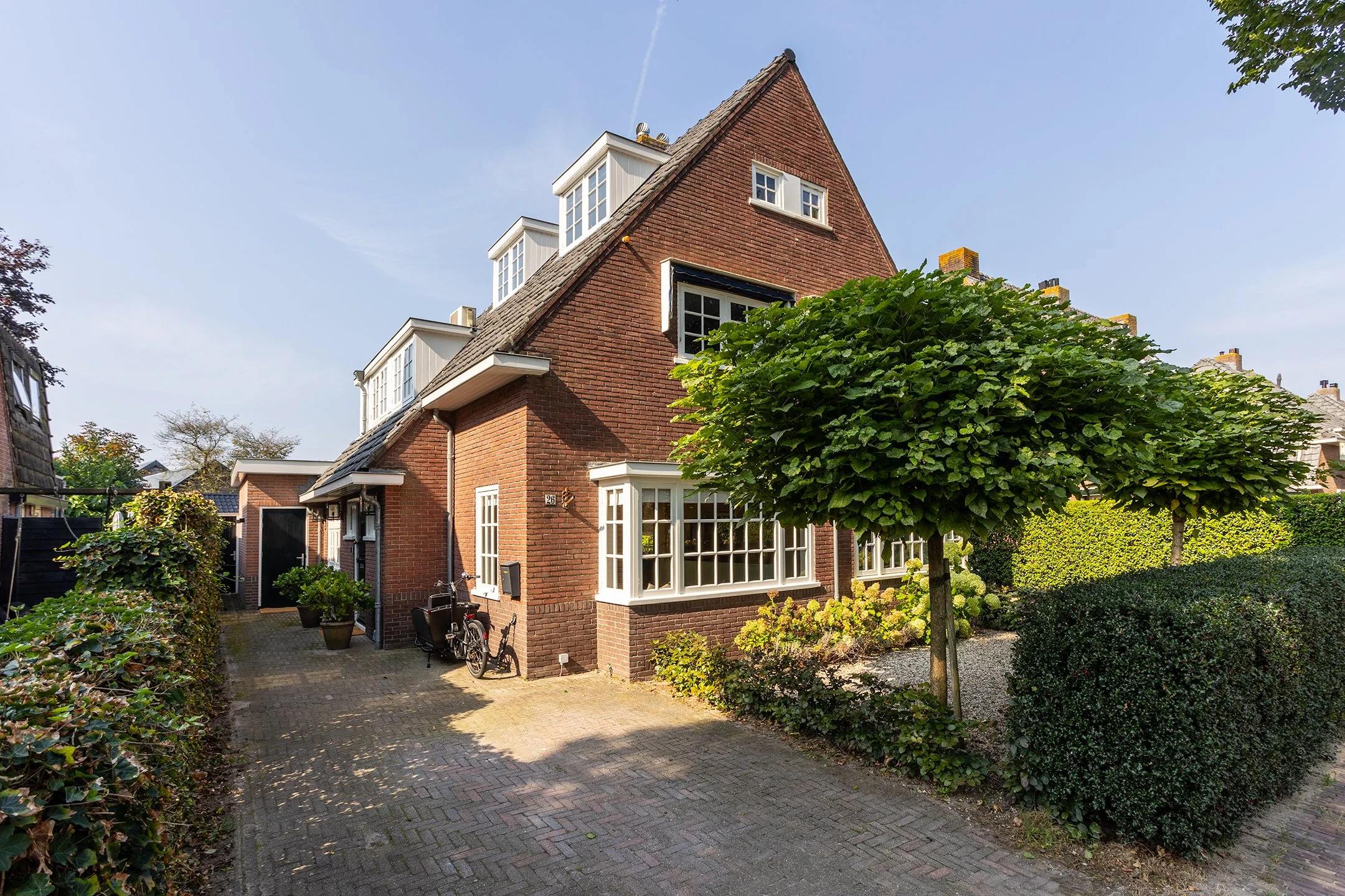 Een rood bakstenen huis met meerdere verdiepingen en witte ramen, omringd door een tuin met een grote groene boom en heggen, onder een blauwe hemel.