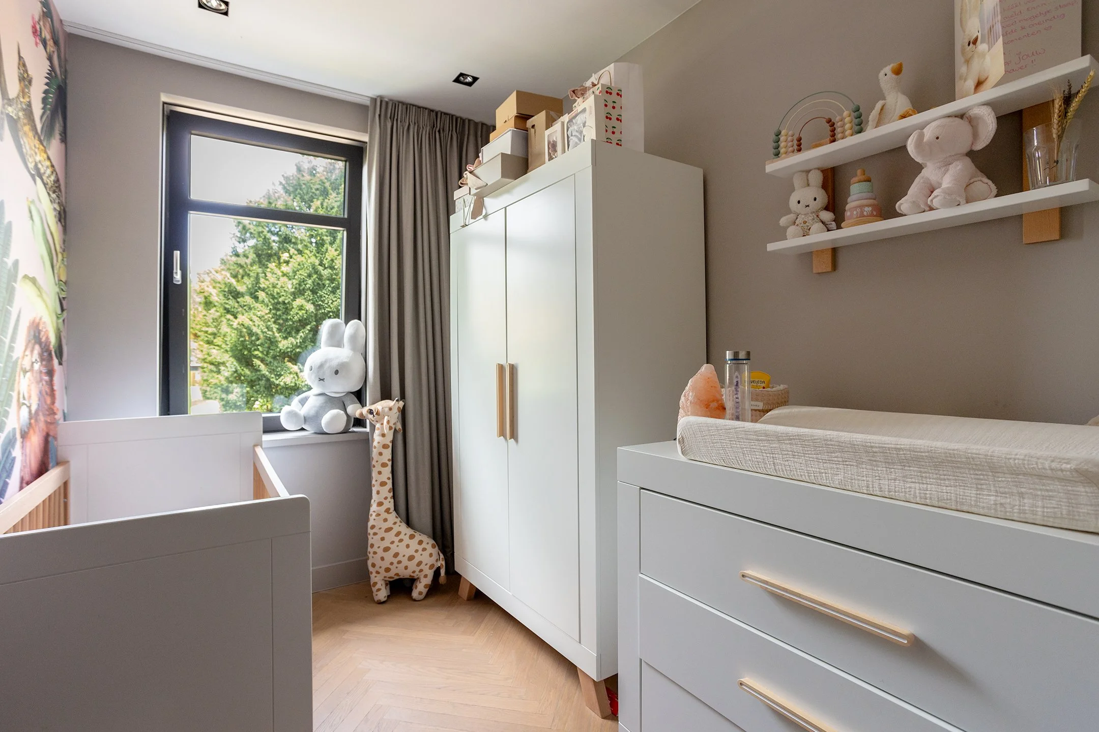 Kinderkamer met witte meubels, speelgoed en knuffels, waaronder een grote knuffelkonijn op de vensterbank, een giraffe knuffel op de vloer, en decoraties op een muurplank.