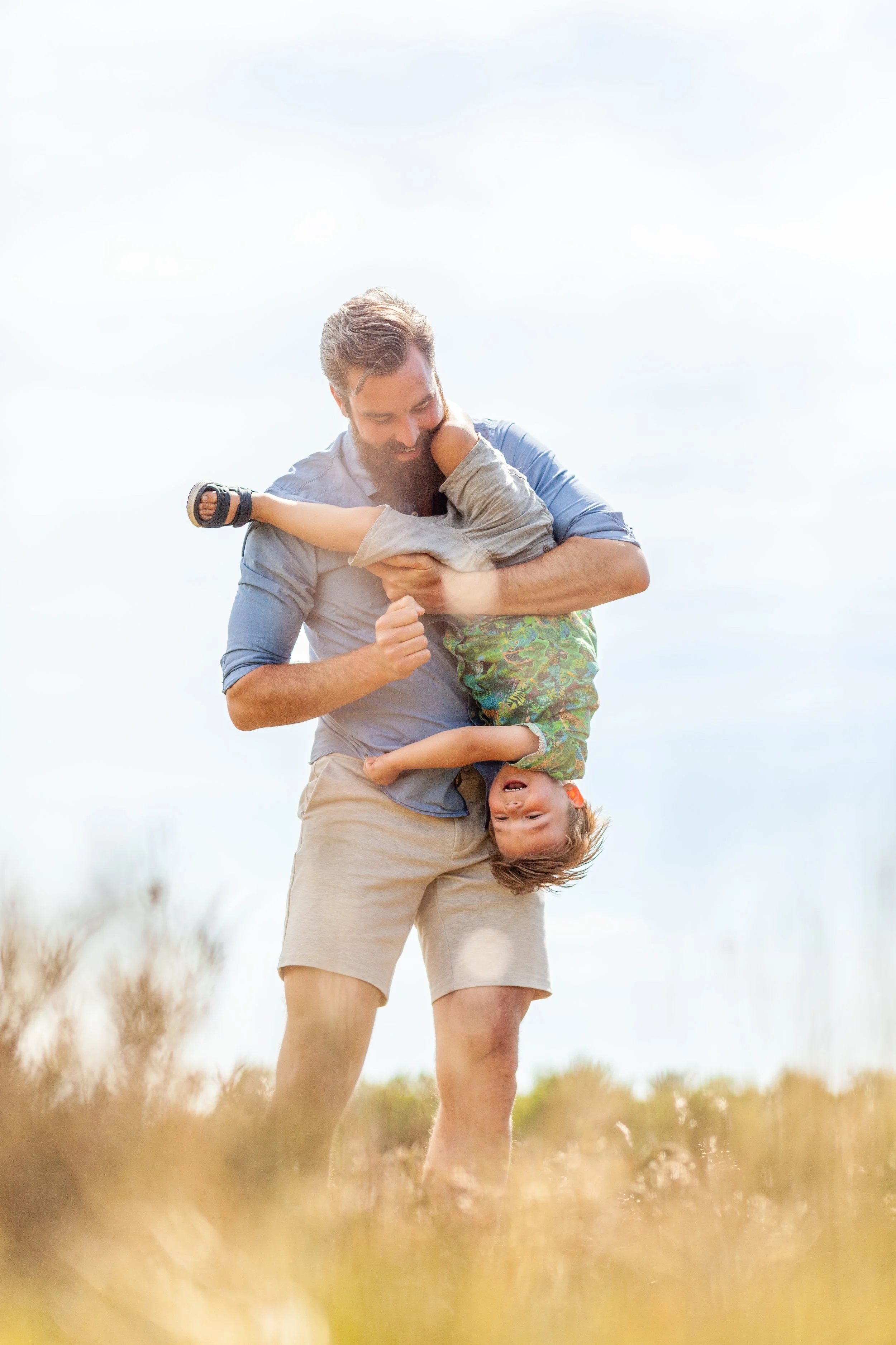 Een man die een jongen vasthoudt terwijl hij lacht, op een open veld onder een lichte hemel.