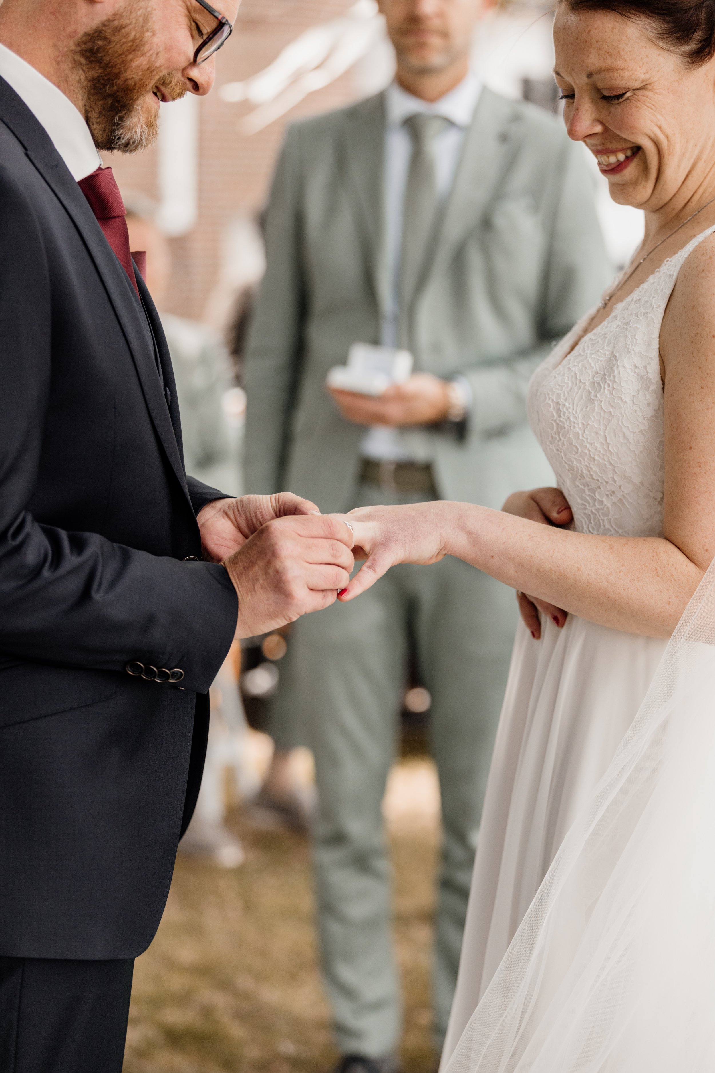 Een stel dat trouwt, de man die de trouwring om de vinger van de vrouw plaatst tijdens de huwelijksceremonie, met een getrouwde vrouw in een witte jurk, en een man in een grijs pak op de achtergrond.