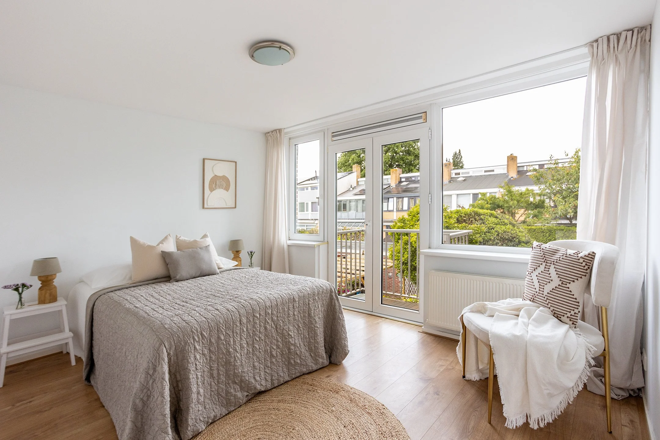 lichtgevelde slaapkamer met groot raam en balkon, beige beddengoed, witte en houten meubels, lichte houten vloer, kunstwerk aan de muur, een stoel met kussen, en lichte gordijnen