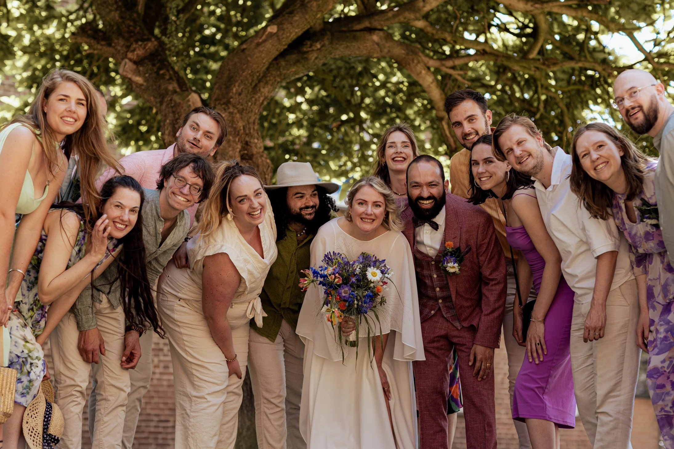 Een groep mensen op een bruiloft, lachend en poserend onder een grote boom, met bloemen en lichte kleding.