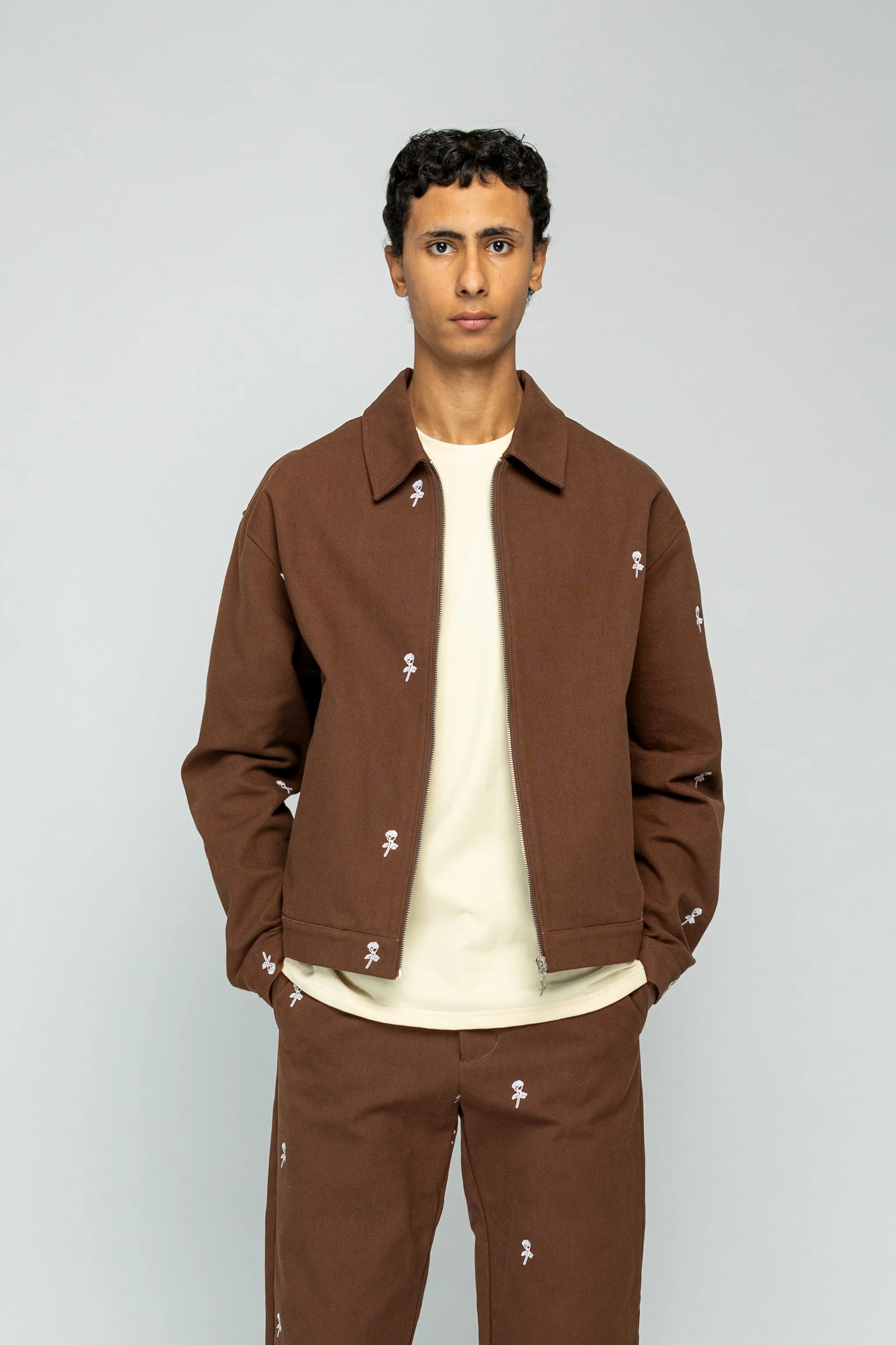 Man in bruin outfit met witte bloemprint, staand voor een lichte achtergrond.