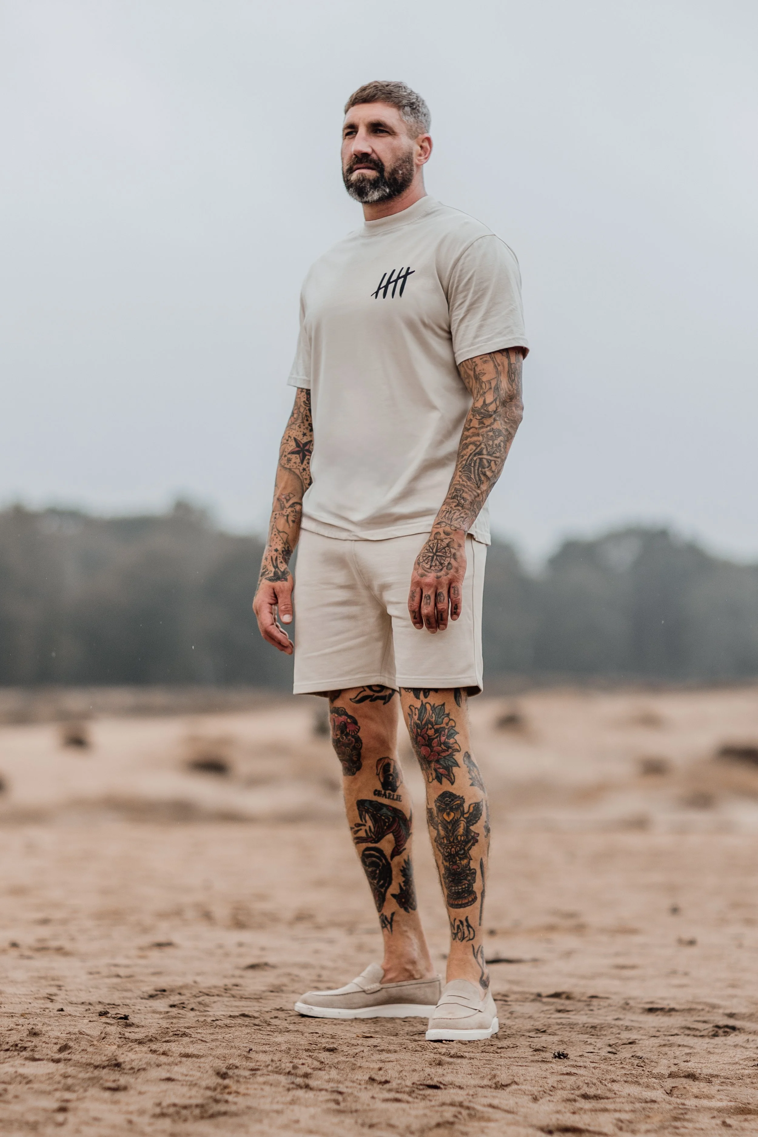 Man met tatoeages staat op een zandige plek, gekleed in een beige t-shirt met een vijf-streep logo en beige korte broek, met lichte schoenen, onder een bewolkte lucht.